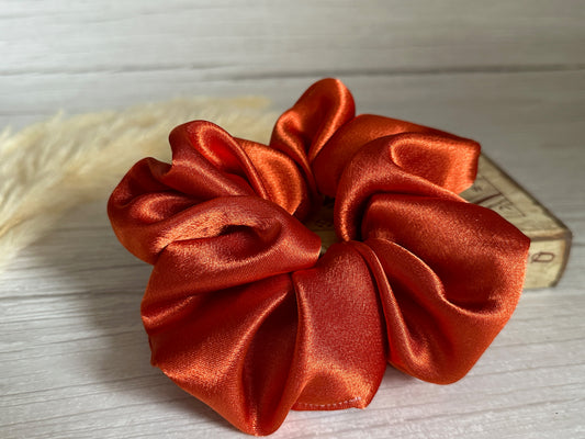 A Silk Extra Large Scrunchie in Burnt Orange from Crafts by Kate rests on a light wooden surface. The handcrafted fabric forms soft, glossy folds, exuding a luxurious sheen. In the background, a small wooden block with numbers is partially visible.