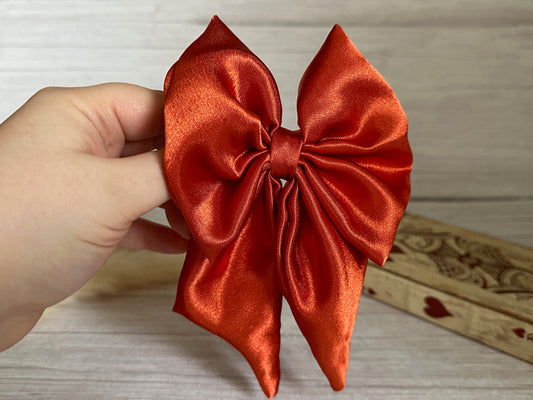 A hand holds a large, shiny burnt orange silk sailor bow clip from Crafts by Kate against a light wooden background. The bow, elegantly tied with two long tails, makes a luxury statement. A handcrafted wooden box adorned with decorative hearts is partially visible in the background.