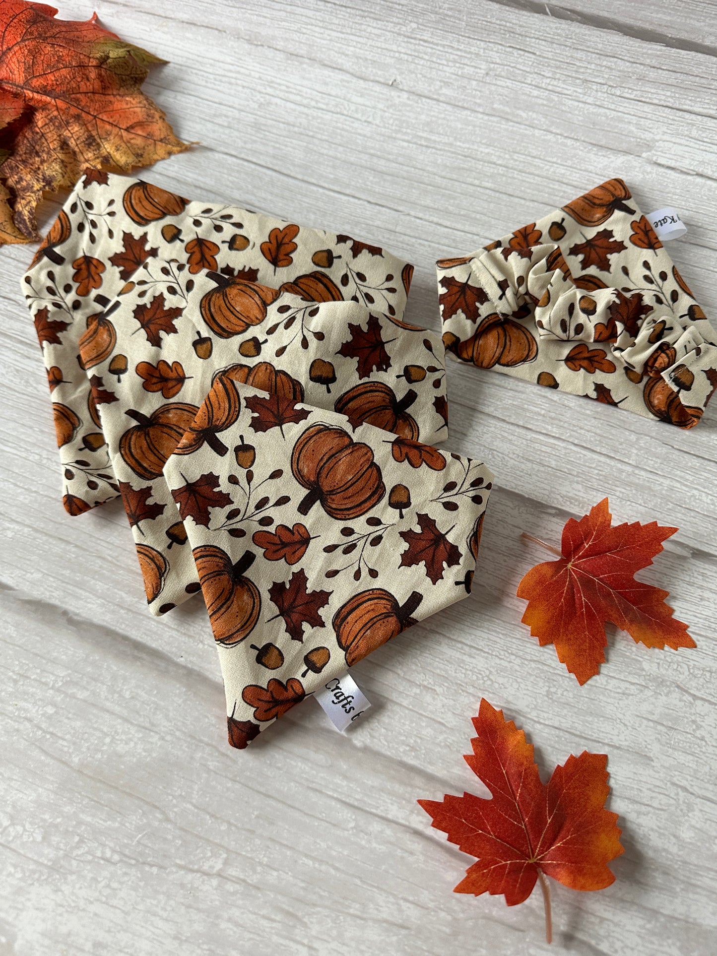 Introducing the "Cotton Pet Scrunchie Bandana - Cottage Core Pumpkins" by Crafts by Kate, a charming autumn-themed fabric set. This collection showcases a delightful motif of pumpkins and leaves elegantly displayed on a beige backdrop. The set includes fabric squares and a bandana featuring similar patterns, artfully accented with three red-orange maple leaves scattered across a light wooden surface.