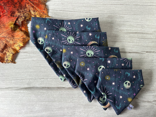 A set of folded Cotton Pet Scrunchie Bandanas - Jack Skeleton from Crafts by Kate, featuring a Halloween theme, laid out on a light wooden surface. The navy blue bandanas are adorned with green alien faces, spaceships, and planets surrounded by stars and bursts. A dried red and orange leaf is placed to the side of the bandanas.