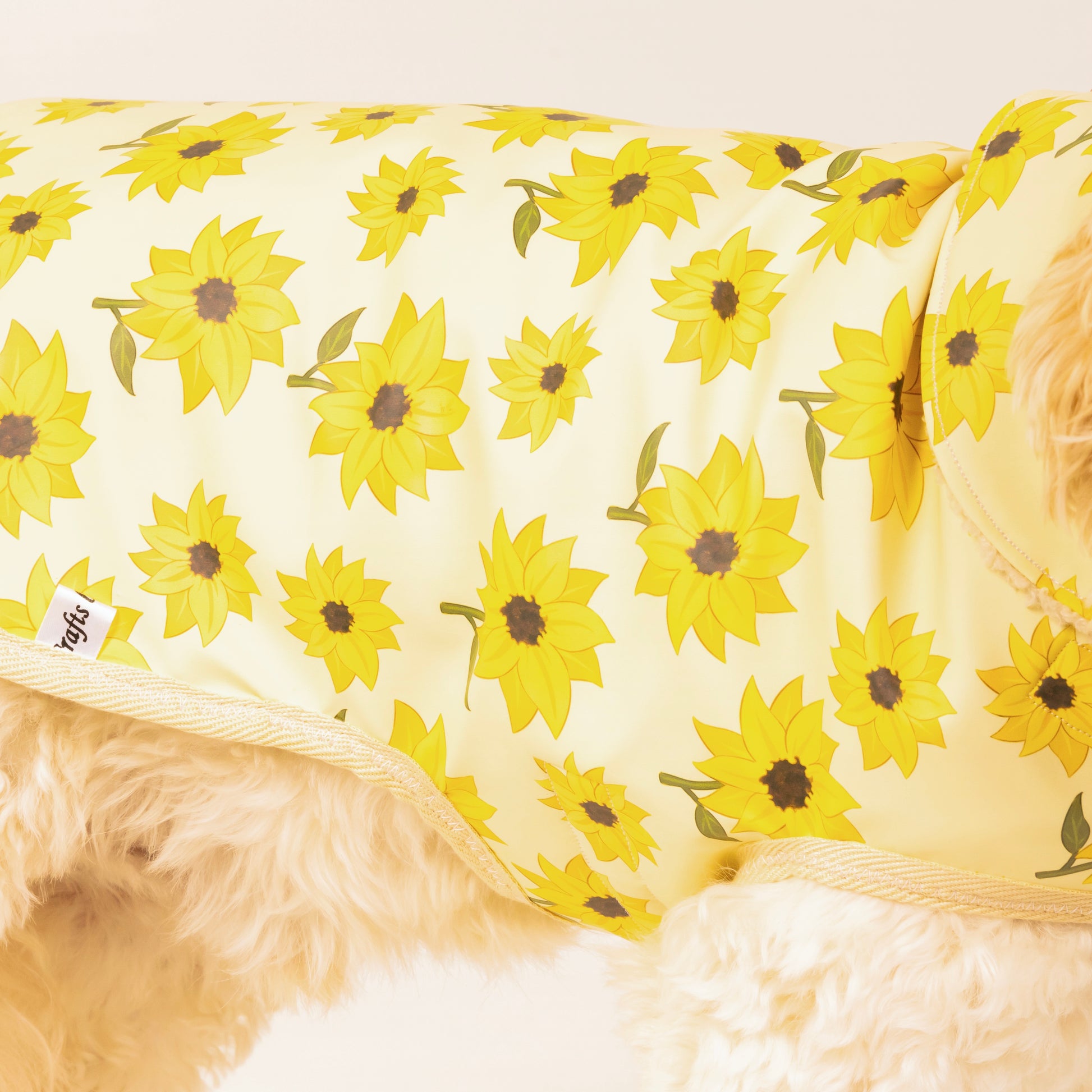 A fluffy dog models the Crafts by Kate Sunflower Fields Pet Raincoat, featuring a vibrant sunflower print and custom harness hole. Only part of the pup is shown, highlighting the coat’s bright details and practical design.