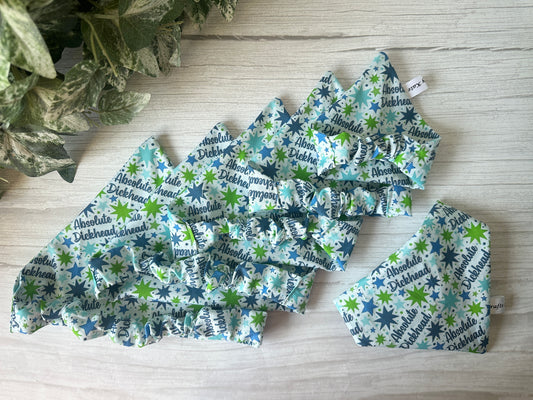 The Cotton Pet Scrunchie Bandana – Absolute Dickhead by Crafts by Kate features blue, green, and white star patterns with bold text, displayed on light wood. Stretchy and stylish, they add flair to your pup’s look. A leafy plant peeks in from the upper left corner.