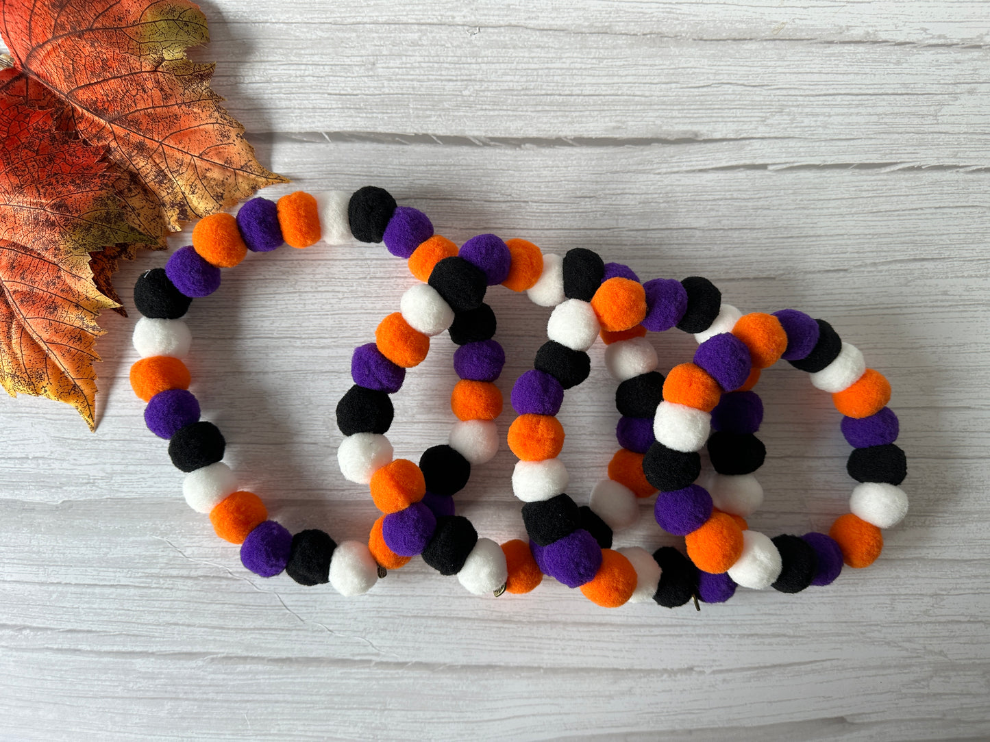 Colorful felt ball garlands made of black, white, orange, and purple balls are arranged in loops on a white wooden surface. A dried autumn leaf is in the top left corner, adding a seasonal touch. Complement this festive decor with the PomPom Pet Collar - Halloween by Crafts by Kate for a complete Halloween look for your furry friend.