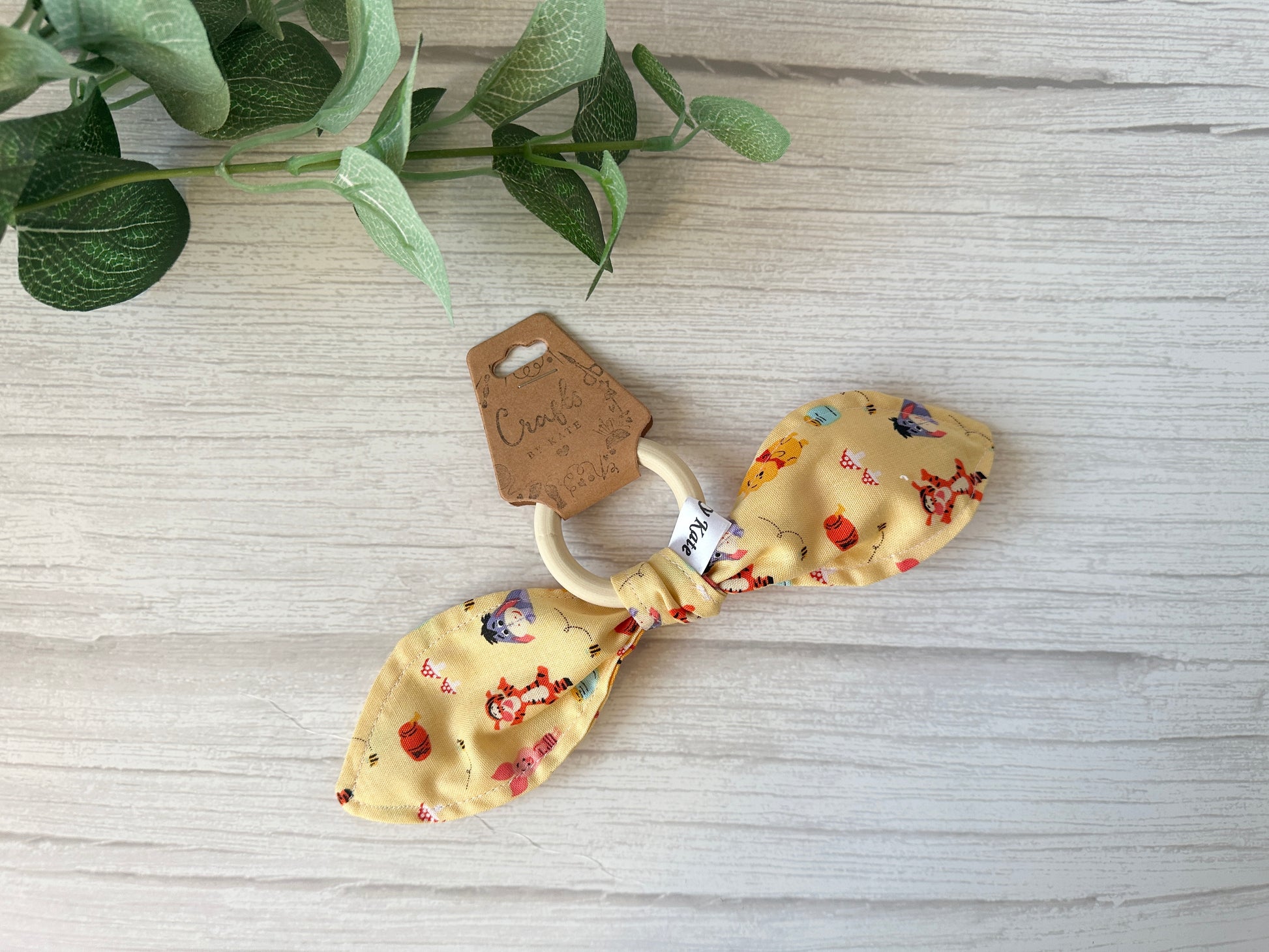A charming baby gift set by Crafts by Kate, featuring a wooden teething ring with ultra-soft cotton in bow form, showcases colorful prints of animals and objects. Comes with a brown cursive tag on a white wooden backdrop with green leaves. Ideal for fans of the Winnie the Pooh & Friends collection!.