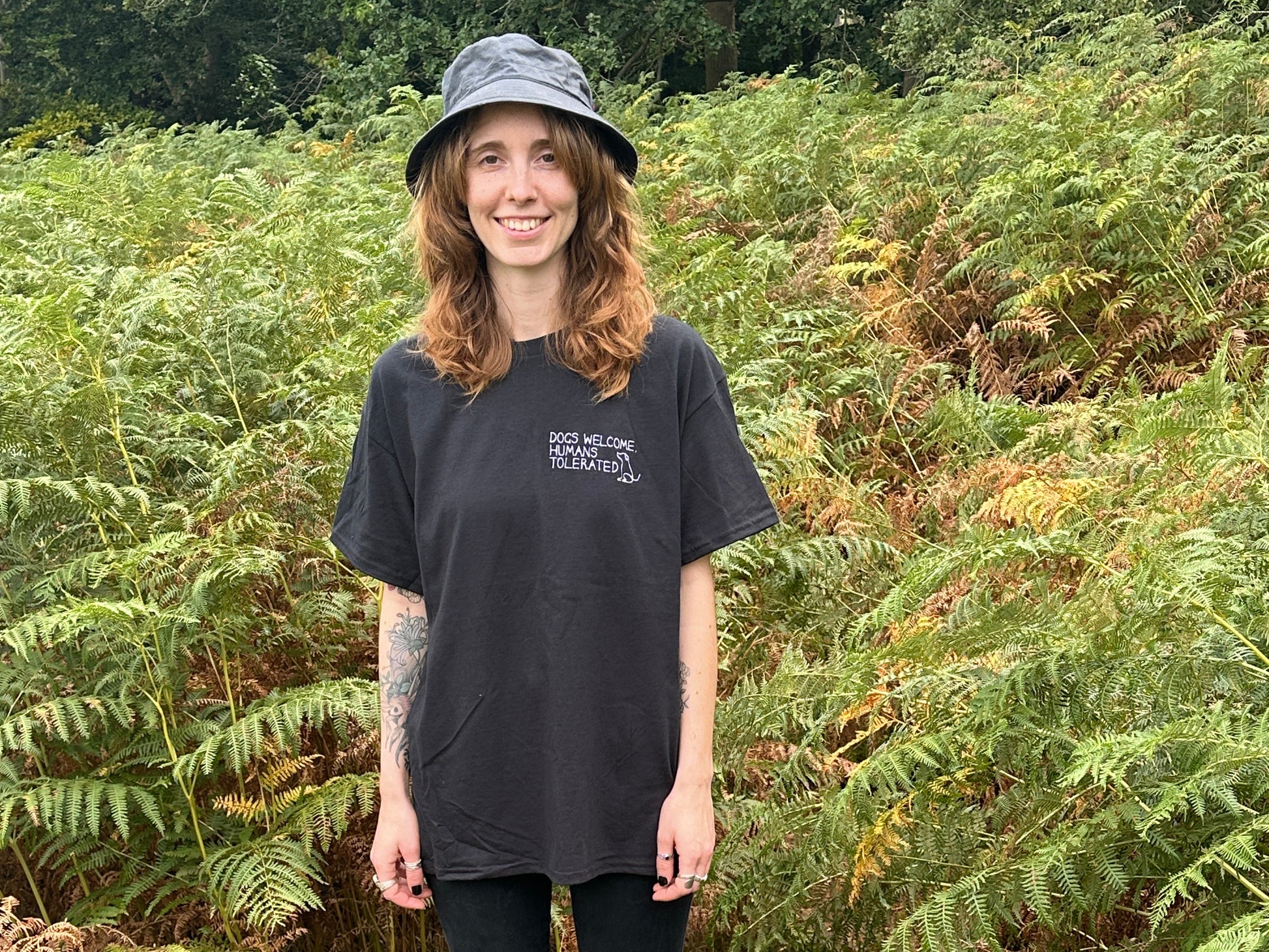 A smiling woman with wavy brown hair wears the Crafts by Kate "Dogs welcome" t-shirt in black and a bucket hat, standing outdoors in front of lush green ferns.
