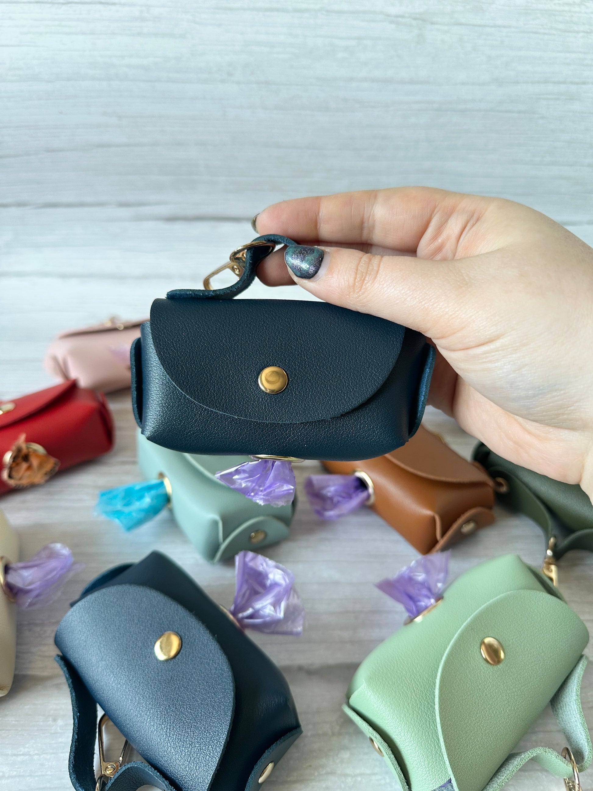 A hand holds a dark teal Crafts by Kate "Pet Boo Bag Holder Handbag" with a gold clasp, evoking chic handbag style. In the background, similar mint green and beige faux leather pouches are scattered, each filled with wrapped purple candies on a light wood surface.