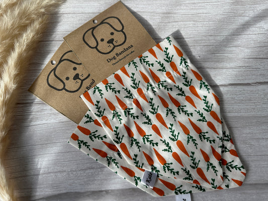 Displayed against a wooden surface are two Cotton Pet Bandanas with collar attachment – Carrots, handcrafted by Crafts by Kate. Each bandana features a white background adorned with an orange carrot pattern. Attached to the bandanas are cardboard tags showcasing a simple dog illustration and the text "Dog Bandana." The collar attachment, made from furry material for easy wear, is partially visible.