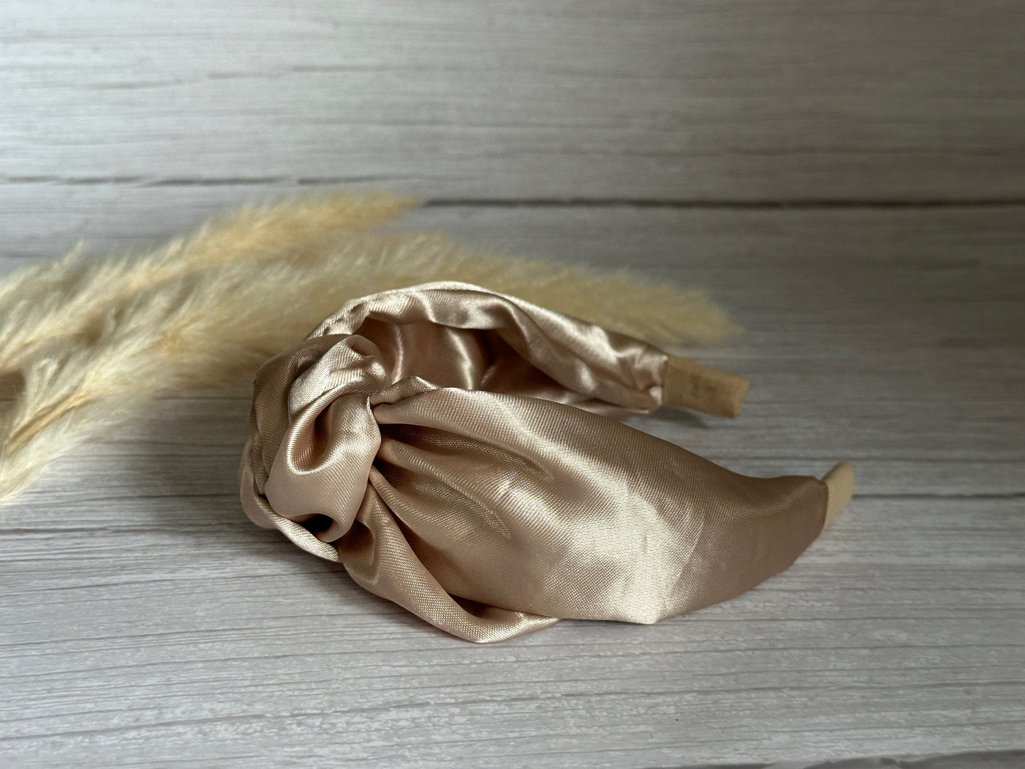 The Silk Knotted Headband - Champagne Gold by Crafts by Kate is elegantly displayed on a light wooden surface, with soft, dried pampas grass in the background enhancing its handcrafted luxury appeal.