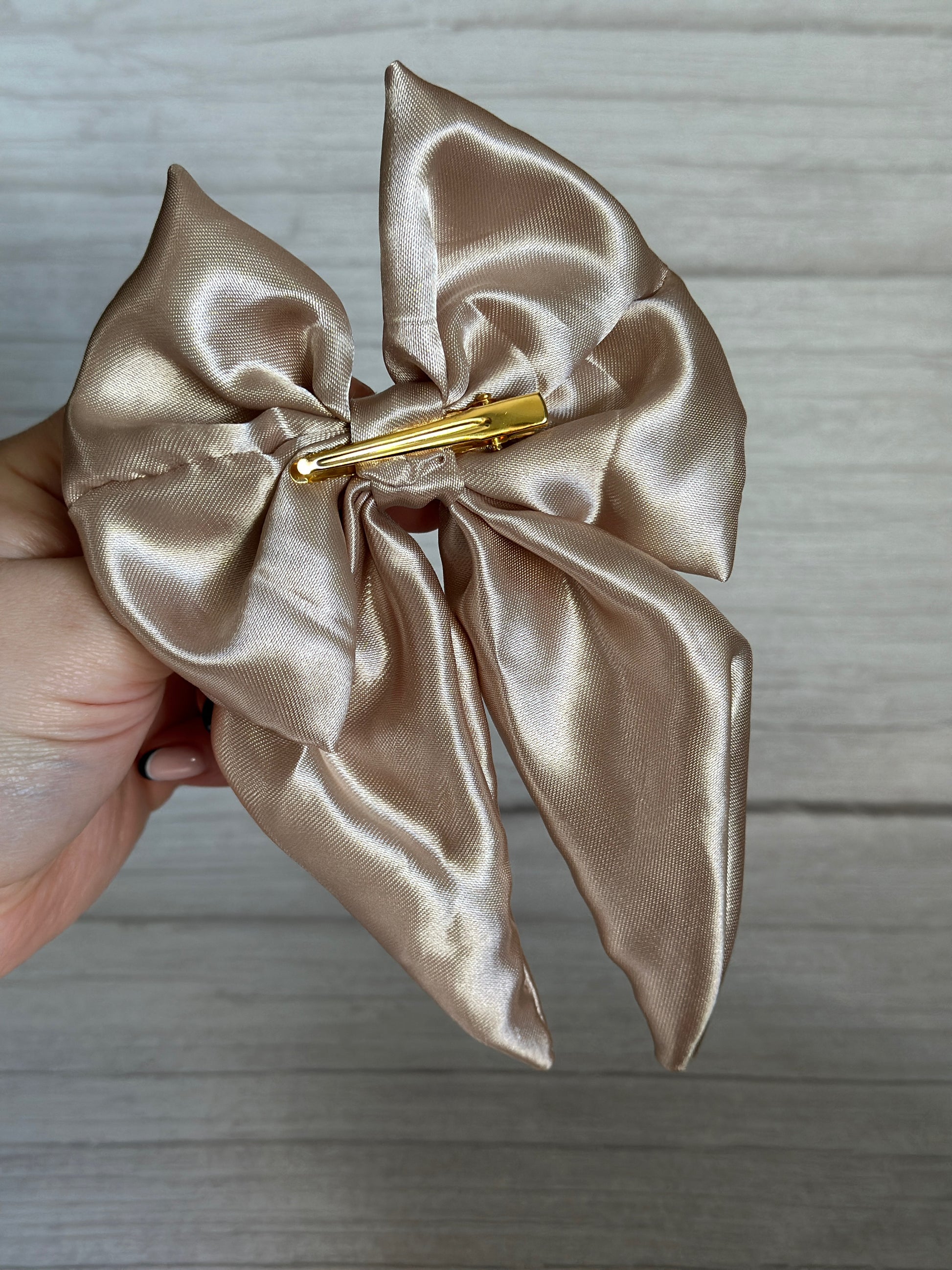 A hand gently holds the Silk Sailor Bow Clip - Champagne Gold by Crafts by Kate, a luxurious piece made from lustrous silk. Fastened with a shining gold barrette clip, it stands out strikingly against the warm wooden backdrop.