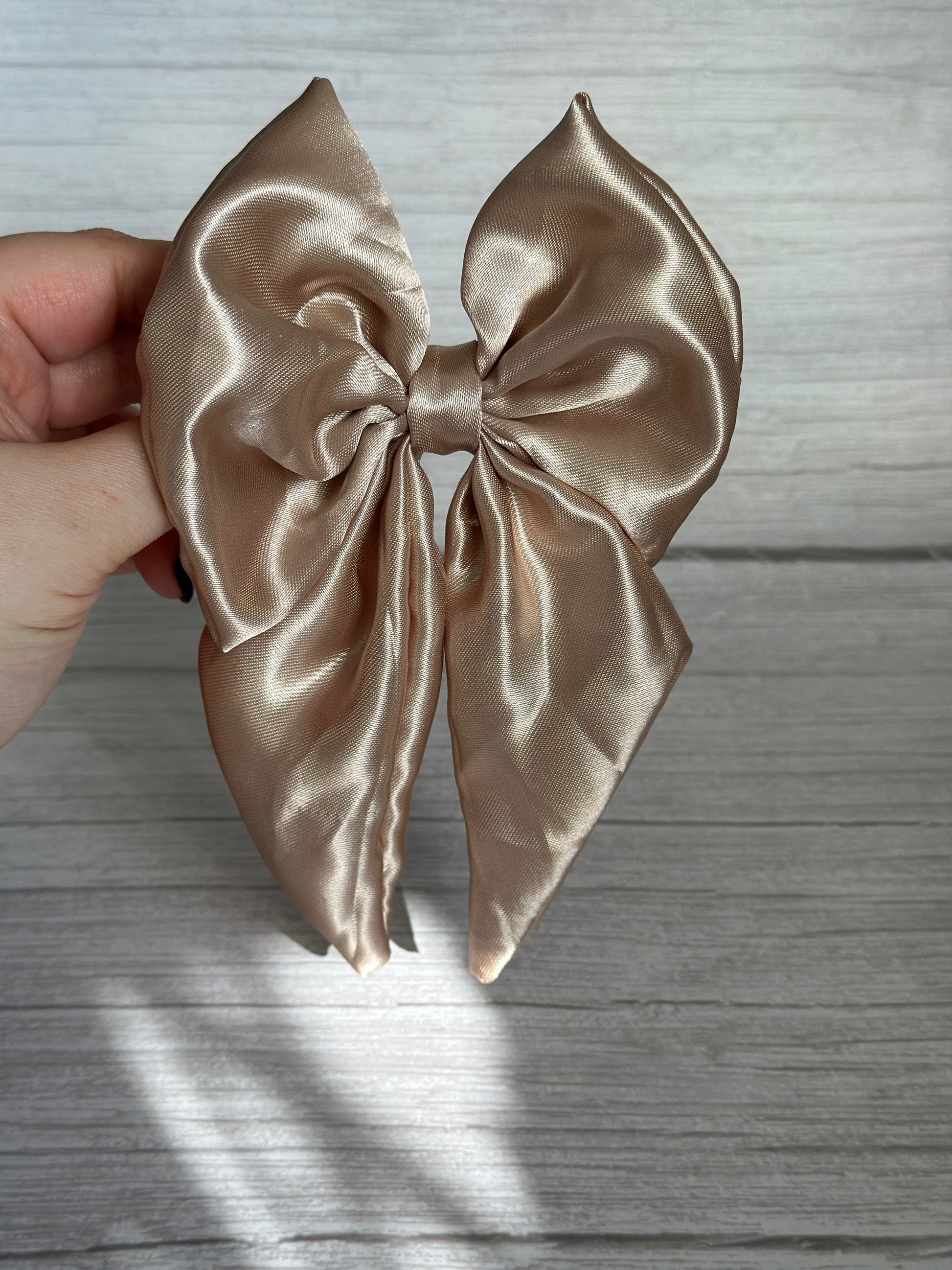 A hand displays the Silk Sailor Bow Clip in champagne gold by Crafts by Kate, crafted from lustrous beige fabric and captured against a light gray wood backdrop.