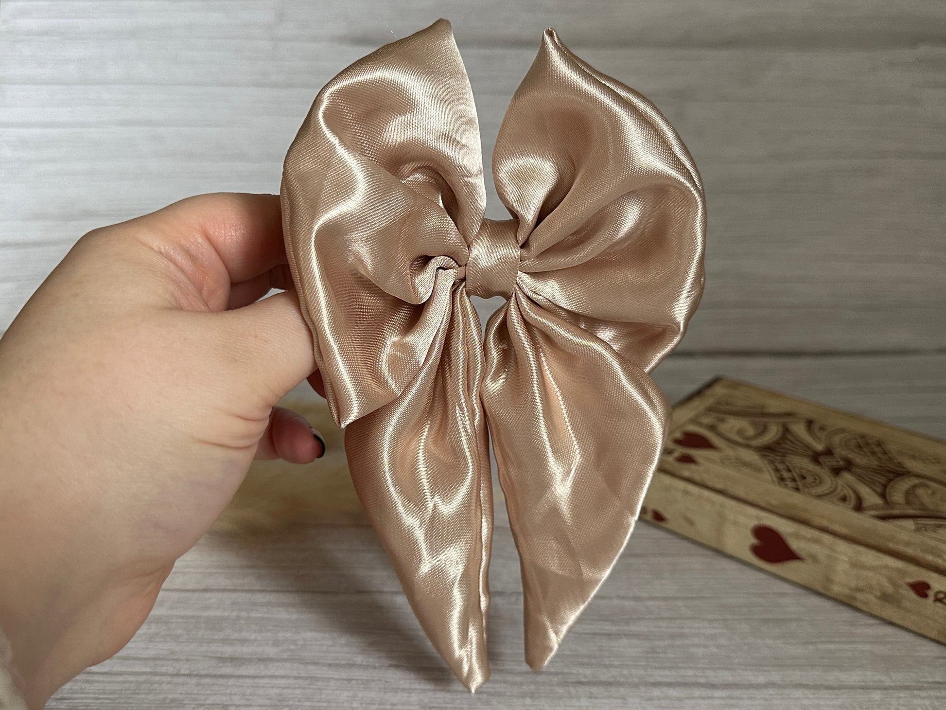 A hand presents a large, shiny Silk Sailor Bow Clip in Champagne Gold from Crafts by Kate against a wooden backdrop. This luxurious statement piece showcases long, elegant loops and tails. To the side, a decorative wooden box adorned with heart designs is partially visible.