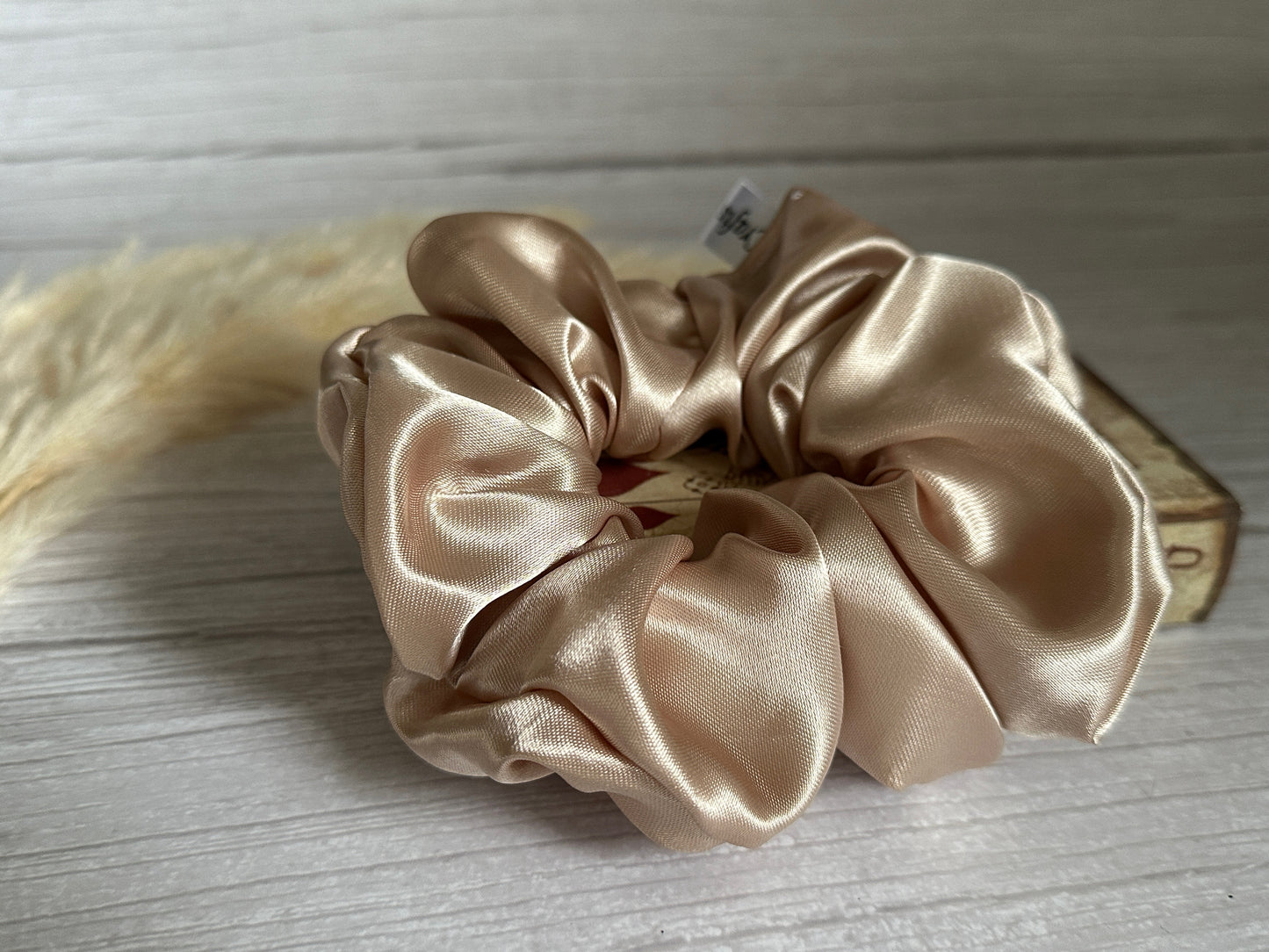 A Silk Extra Large Scrunchie in Champagne Gold from Crafts by Kate is elegantly displayed on a wooden surface, accented by delicate pampas grass on the left, all set against a light backdrop.