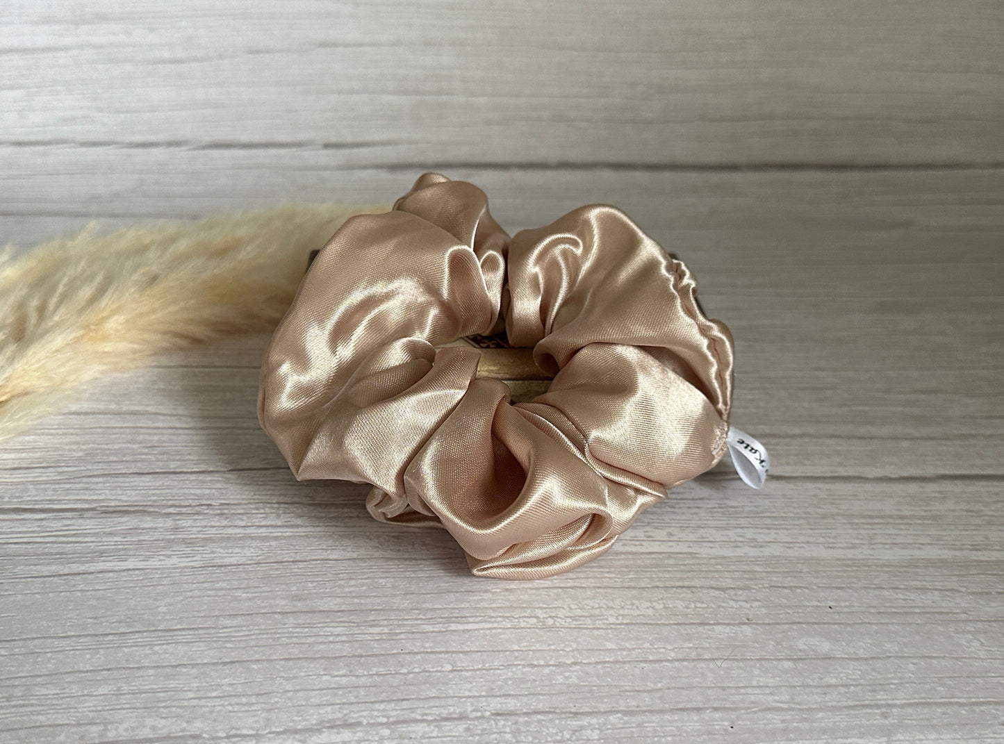 A Silk Extra Large Scrunchie in Champagne Gold by Crafts by Kate rests elegantly on a light wooden surface, complete with a small white tag. Fluffy, cream-colored dried grasses are partially visible on the left.