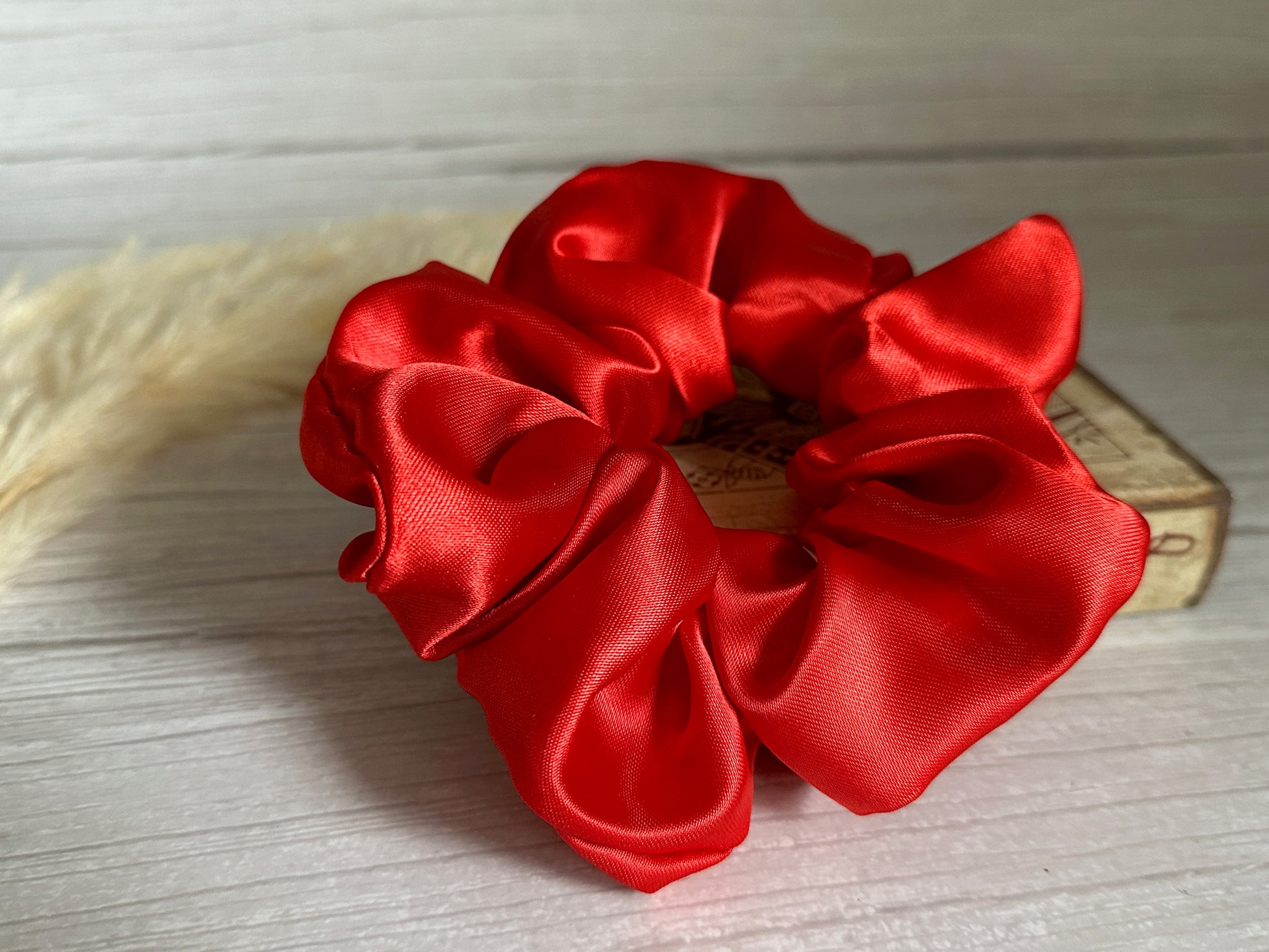 A Silk Extra Large Scrunchie in Cherry Red from Crafts by Kate rests on a light wooden surface. Behind it, a decorative rectangular box is partially visible, highlighting its handcrafted elegance, while a soft, beige pampas grass stalk gracefully stands to the left.