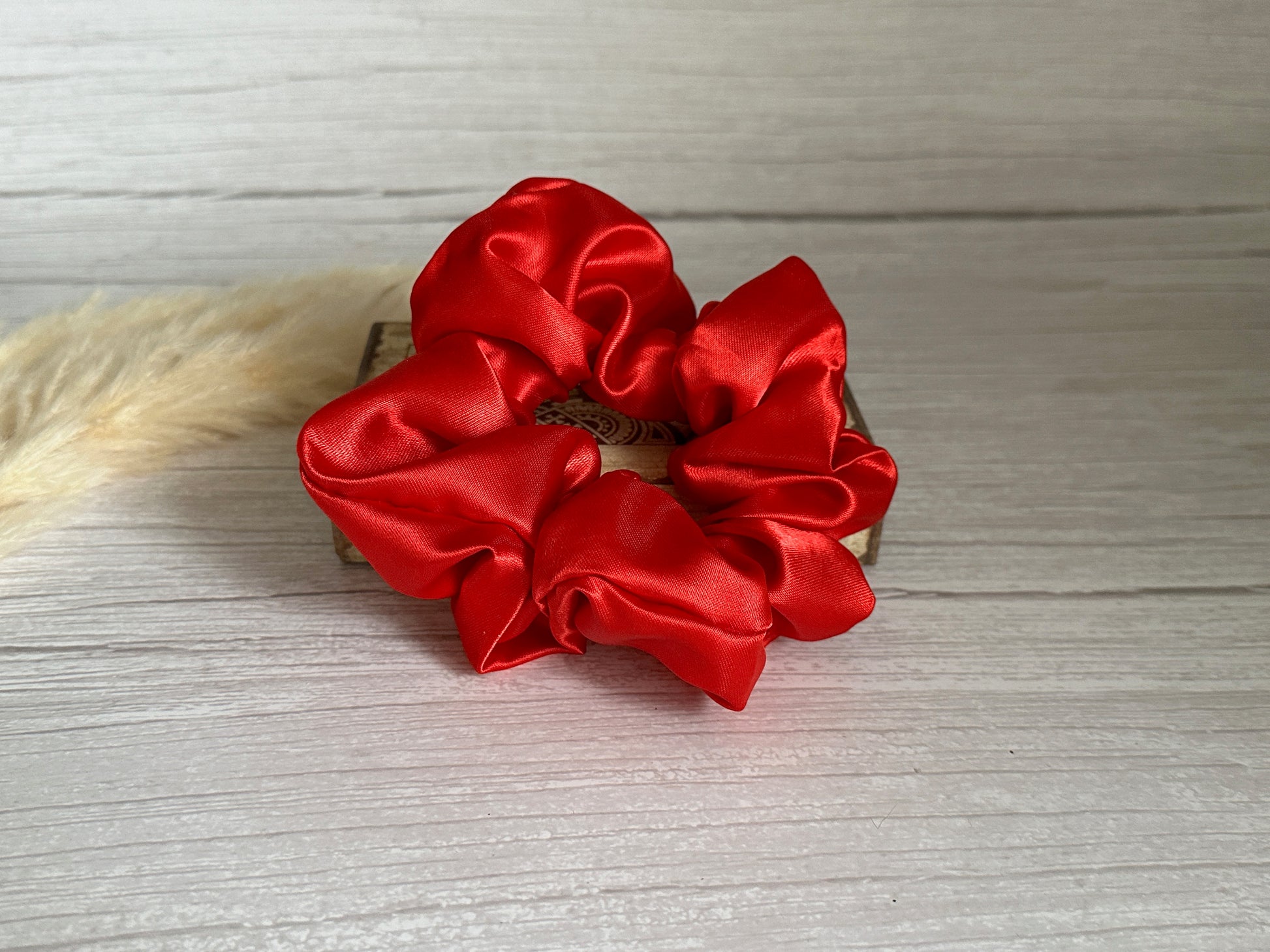 A Silk Extra Large Scrunchie in Cherry Red from Crafts by Kate is elegantly displayed on a wooden surface. The handcrafted scrunchie highlights its glossy texture, while a light beige pampas grass stem peeks in from the left side of the image.