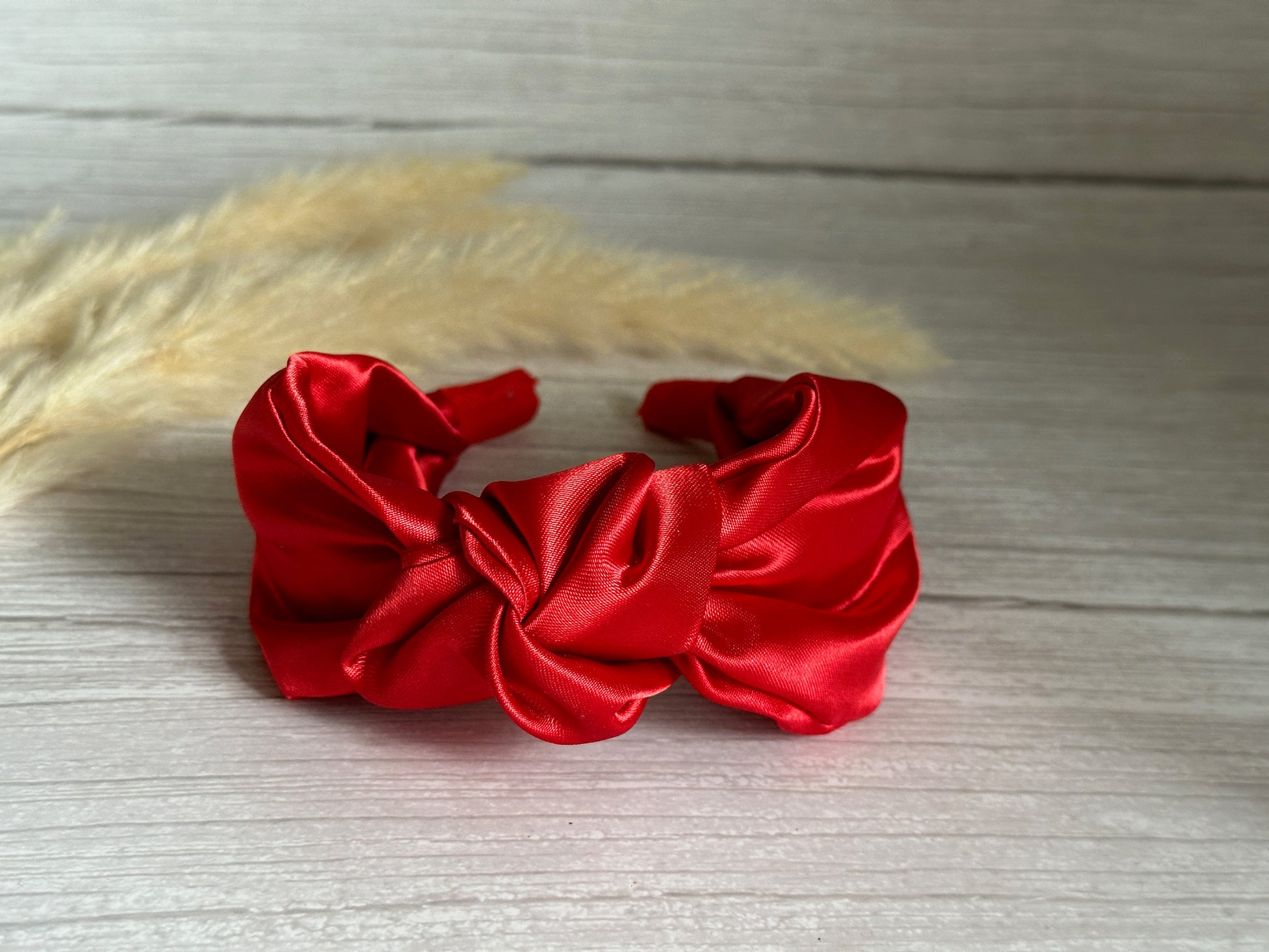 The Silk Knotted Headband in Cherry Red by Crafts by Kate, epitomizing handcrafted luxury, rests gracefully on a light wood surface. Behind it, soft beige pampas grass plumes provide a gentle backdrop.