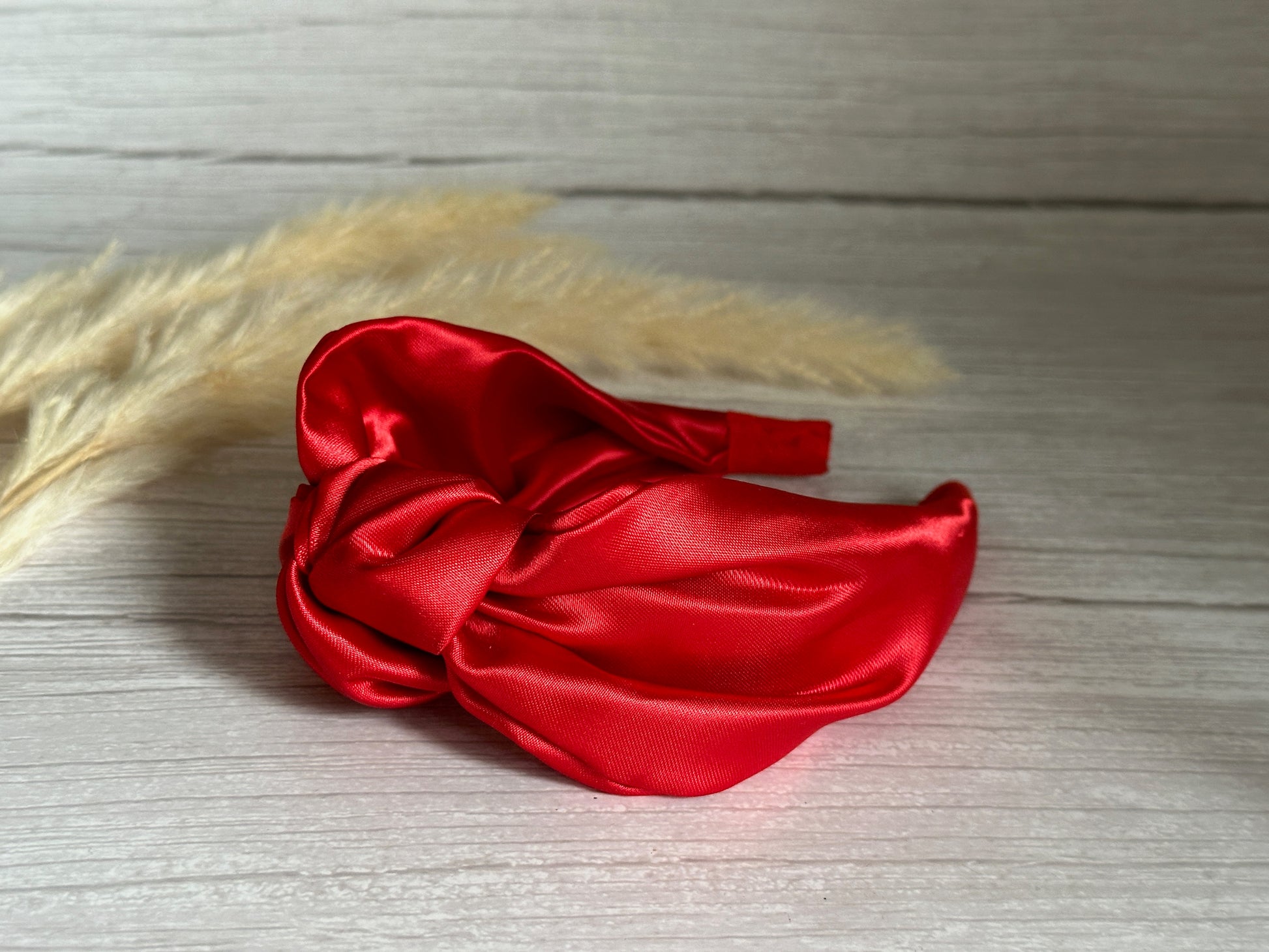 The Silk Knotted Headband - Cherry Red by Crafts by Kate, featuring a vibrant design in luxurious satin and a knot detail, is elegantly placed on a light wood surface. Beige pampas grass in the background complements the scene with its soft, natural touch while offering long wear comfort.