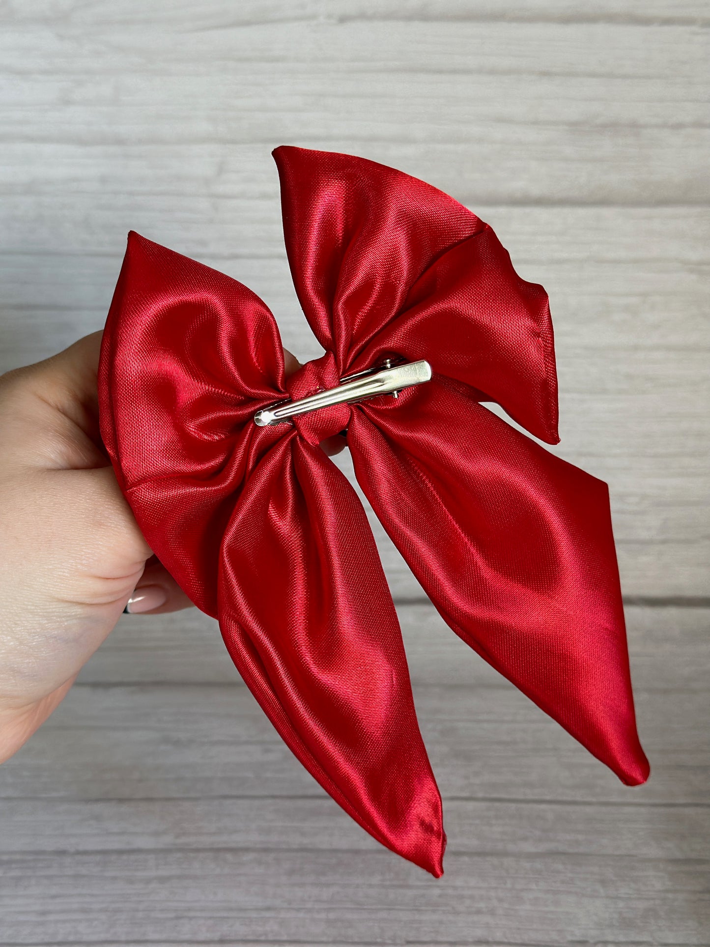 A hand holds the Silk Sailor Bow Clip in cherry red from Crafts by Kate, featuring an elegant silk design with a secure crocodile clip attachment on the back. The vibrant accessory stands out beautifully against the wooden background.