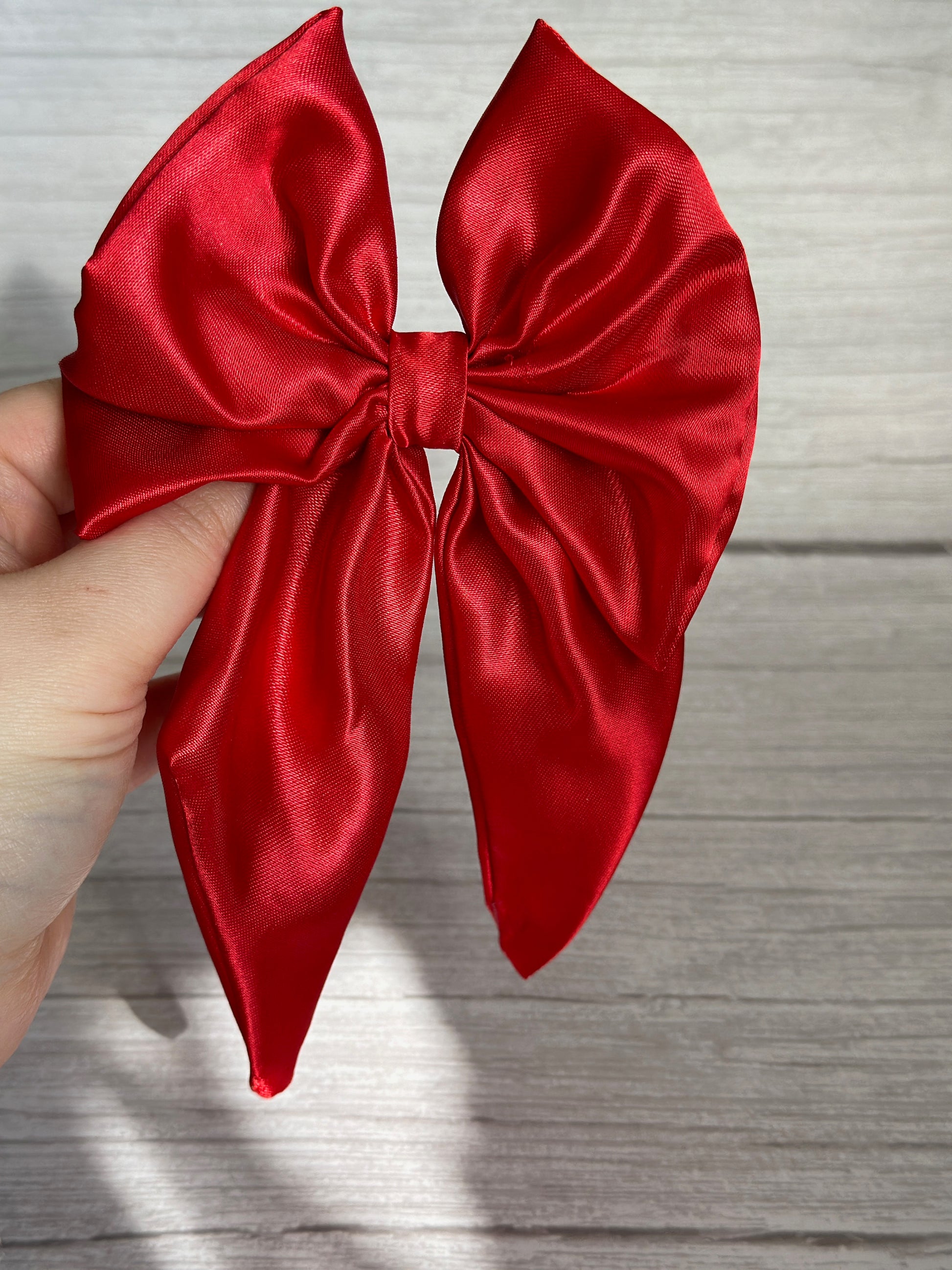 A hand holds the Crafts by Kate Silk Sailor Bow Clip in cherry red over a light woodgrain background. The satin's glossy finish shines brilliantly in the sunlight, emphasizing its exquisite texture and large loops.