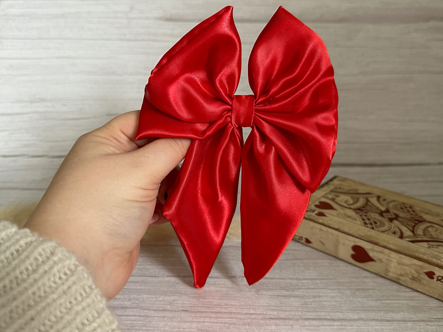 A hand holds the elegant Silk Sailor Bow Clip - Cherry Red from Crafts by Kate against a light wooden background. The bow, with its luxurious cherry red silk, is the centerpiece, gleaming beautifully. To the right, part of a decorative wooden box adorned with heart patterns adds to the sophisticated scene.