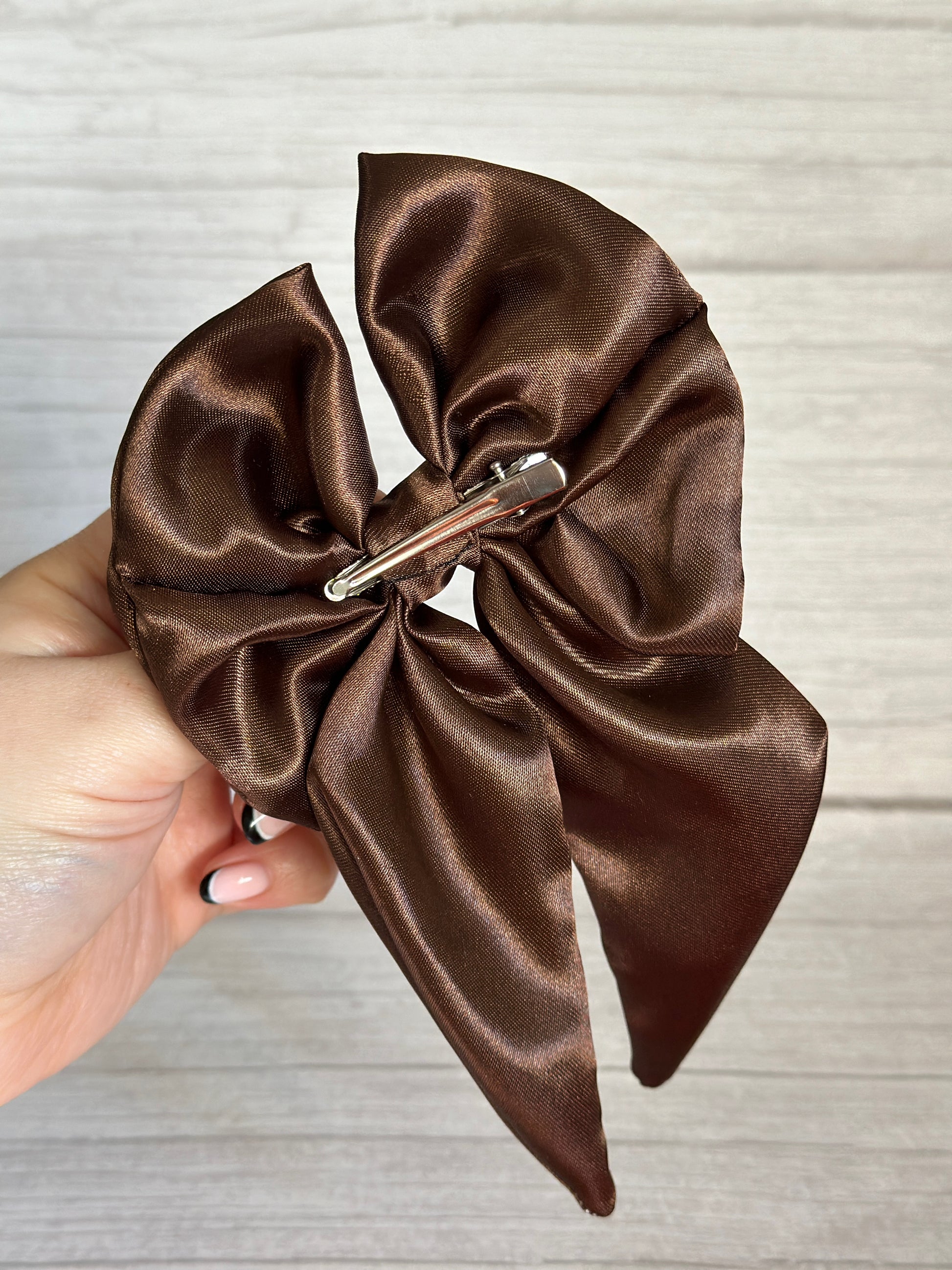 A hand delicately holds the Silk Sailor Bow Clip in chestnut brown, crafted by Crafts by Kate. This luxurious bow, with its shiny silk texture and a sturdy metal clip on the back, is gracefully positioned against a light wooden background.