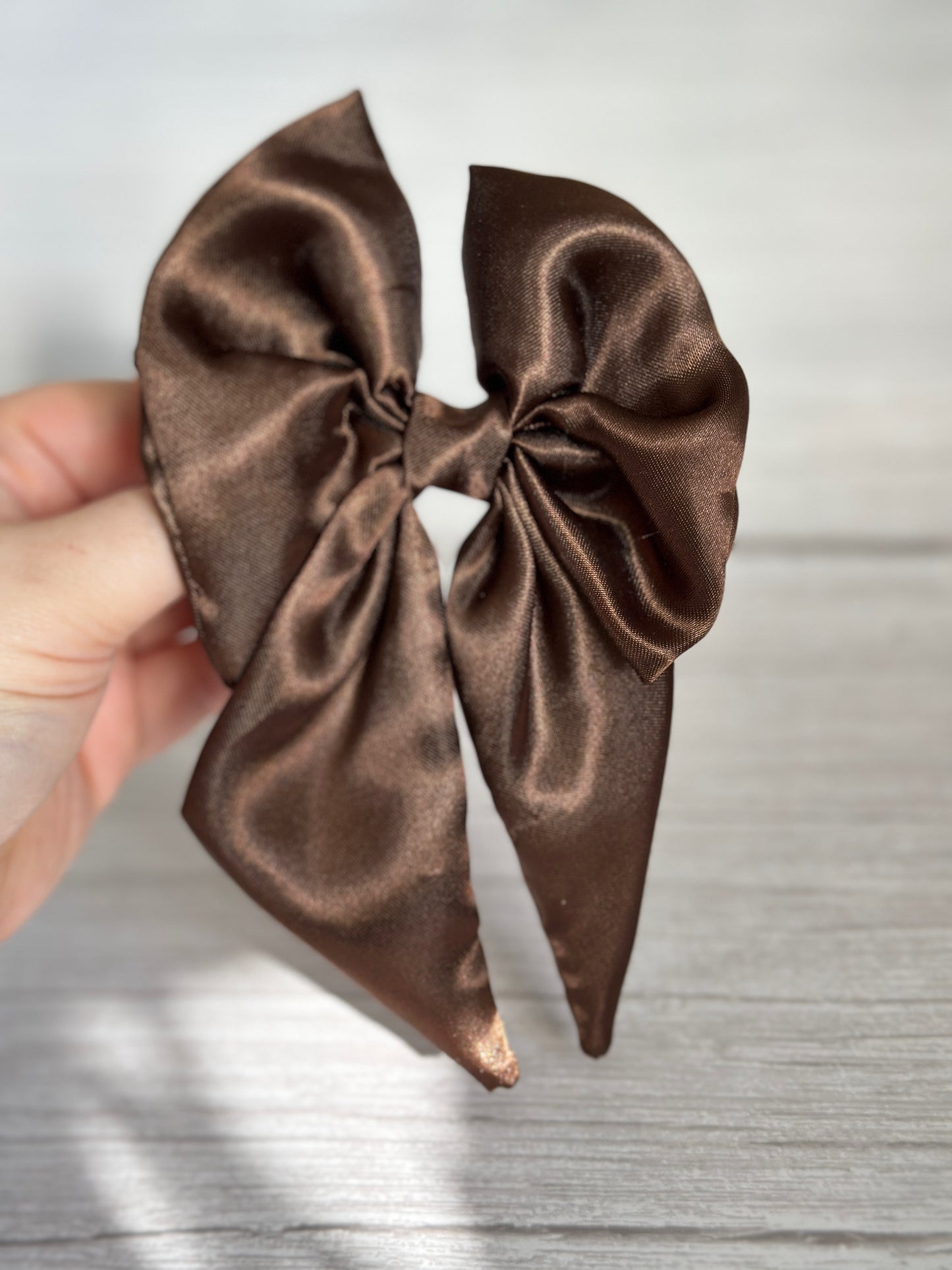 A hand elegantly holds the luxurious Silk Sailor Bow Clip in Chestnut Brown by Crafts by Kate against a light gray wooden surface. The bow is tied neatly with long tails draping gracefully down.