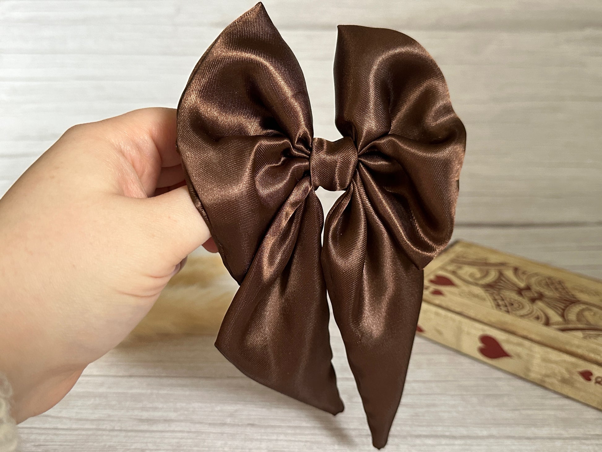 A hand gracefully holds the opulent Silk Sailor Bow Clip in Chestnut Brown from Crafts by Kate against a light wooden background. The sumptuous gloss of the clip enhances its magnificence, while a decorative box adorned with heart designs is partially visible in the background, adding an extra touch of charm.