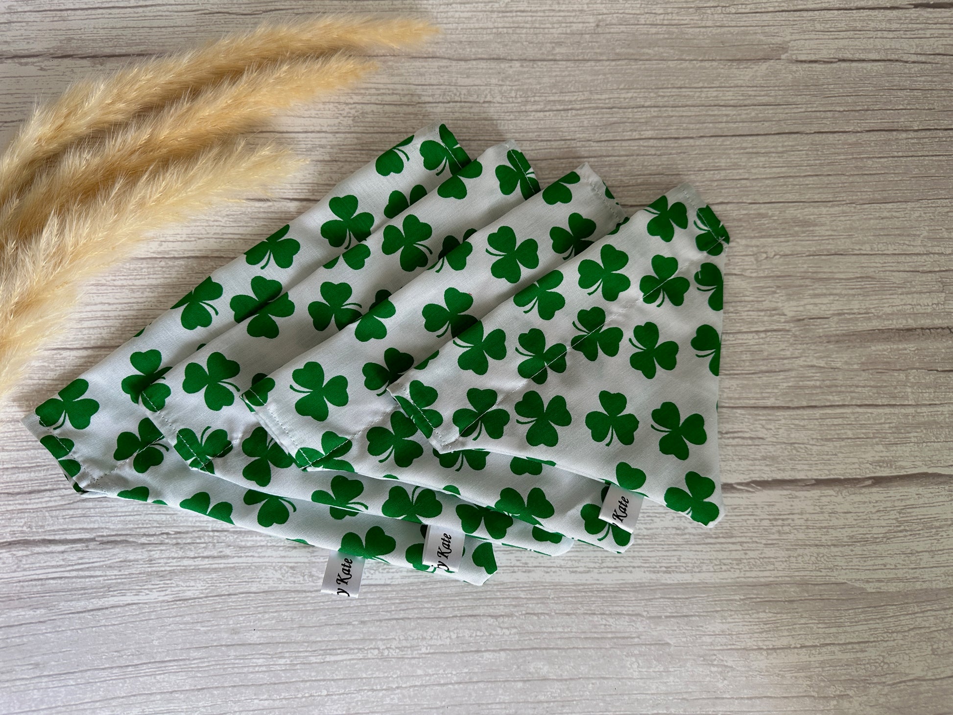 Four white cloth napkins patterned with green four-leaf clovers are neatly arranged in a fan shape on a light wooden surface. Some beige dried grasses are placed to the left of the napkins, adding a touch of natural decor. The same clover fabric from Crafts by Kate's Cotton Pet Bandana with collar attachment could also be used for these handcrafted cotton items.