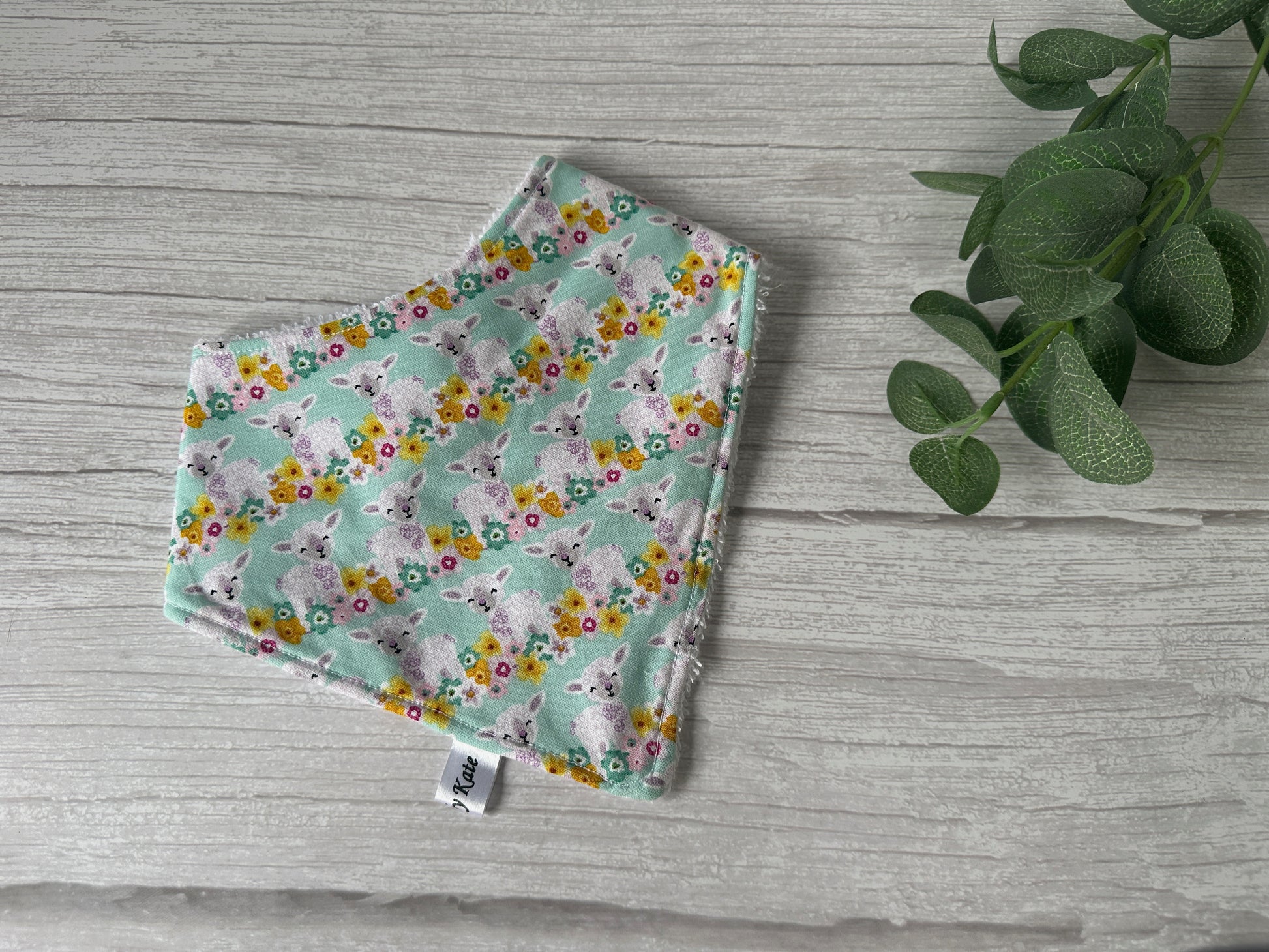 A Cotton & Terry Towelling Baby Bandana Bib - Spring Lambs by Crafts by Kate is laid flat on a light wooden surface, showcasing its pastel green background and adorable lamb and flower pattern. A sprig of green leaves is placed to the right of the handcrafted bib for decoration.