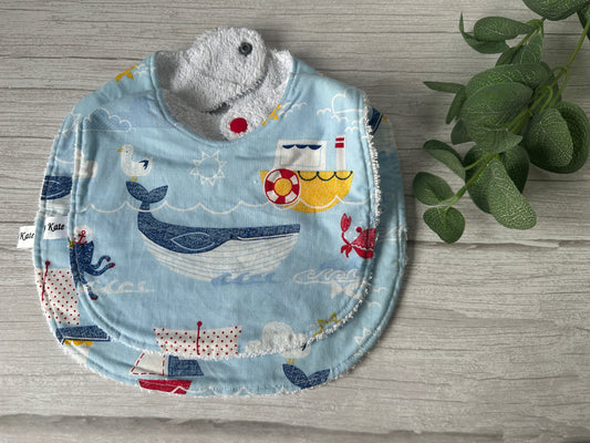 The "Boat Adventures" Cotton & Terry Towelling Baby Traditional Bib by Crafts by Kate, featuring nautical-themed illustrations of boats, a whale, an anchor, and a crab, is laid flat on a light wooden surface. The handcrafted ultra-soft cotton fabric appears soft and absorbent with a snap closure at the neck. A green leafy plant is partially visible to the right.