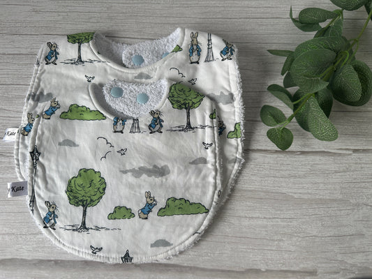 The Cotton & Terry Towelling Baby Traditional Bib - Peter Rabbit's Adventures by Crafts by Kate is a handcrafted, white baby bib adorned with charming cartoon illustrations of trees, birds, clouds, and clothing-clad bunnies. It features two blue snap buttons for easy fastening. A small plant with green leaves is placed next to the cotton bib on a light wooden surface.