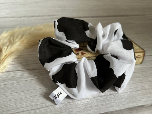 A luxurious Cotton Extra Large Scrunchie - Cow Print by Crafts by Kate rests on a light wooden surface, with a small piece of pampas grass in the background. This handcrafted cotton accessory features an elegant tag labeled "Katie," adding a touch of personalized charm.