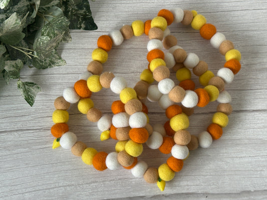 Three garlands of white, yellow, orange, and light brown felt balls rest on light wood beside green leaves—perfect inspiration for the PomPom Pet Collar - Rubber Ducks by Crafts by Kate.