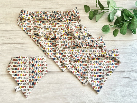 Crafts by Kate presents the Cotton Pet Scrunchie Bandana - Gotcha Day, featuring colorful letters on Barkday fabric. This collection includes bandanas, collars, and bow ties displayed on a light wooden surface beside green leaves.