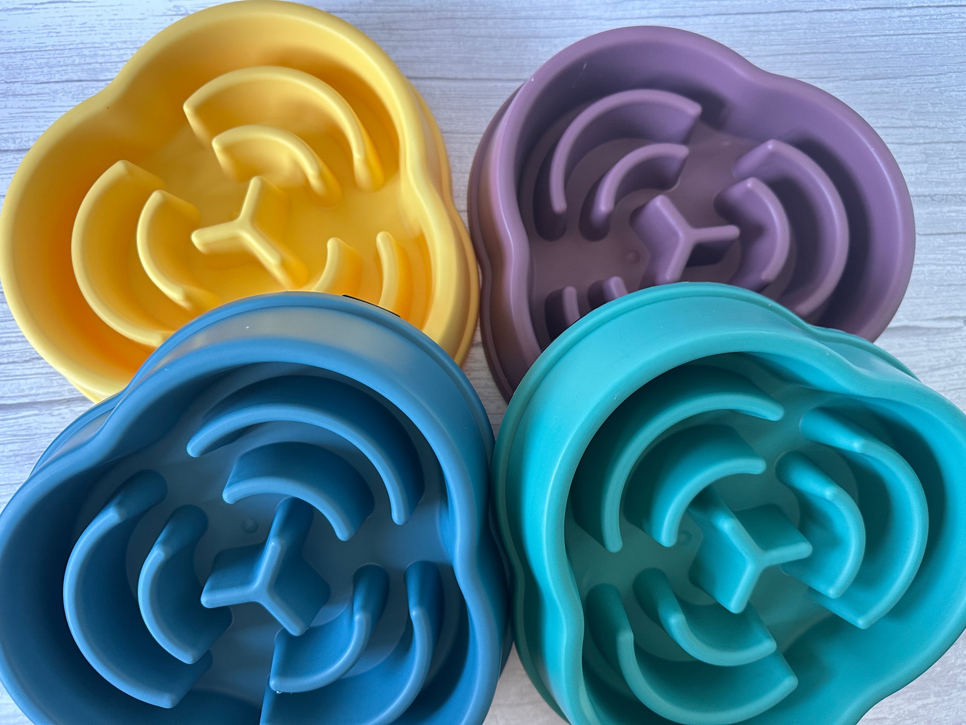 Four vibrant Crafts by Kate Slow Feeder Pet Bowls in yellow, purple, blue, and teal are elegantly placed on a white surface. These flower-shaped bowls have grooved patterns to encourage slow eating for improved digestion, adding a splash of color to mealtime while promoting healthier pet eating habits.