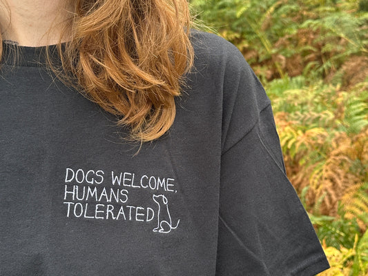 A person with long, light brown hair wears a black oversized unisex "Dogs welcome t-shirt" by Crafts by Kate, featuring white text "DOGS WELCOME, HUMANS TOLERATED" and a simple dog outline. Green foliage is visible in the background.