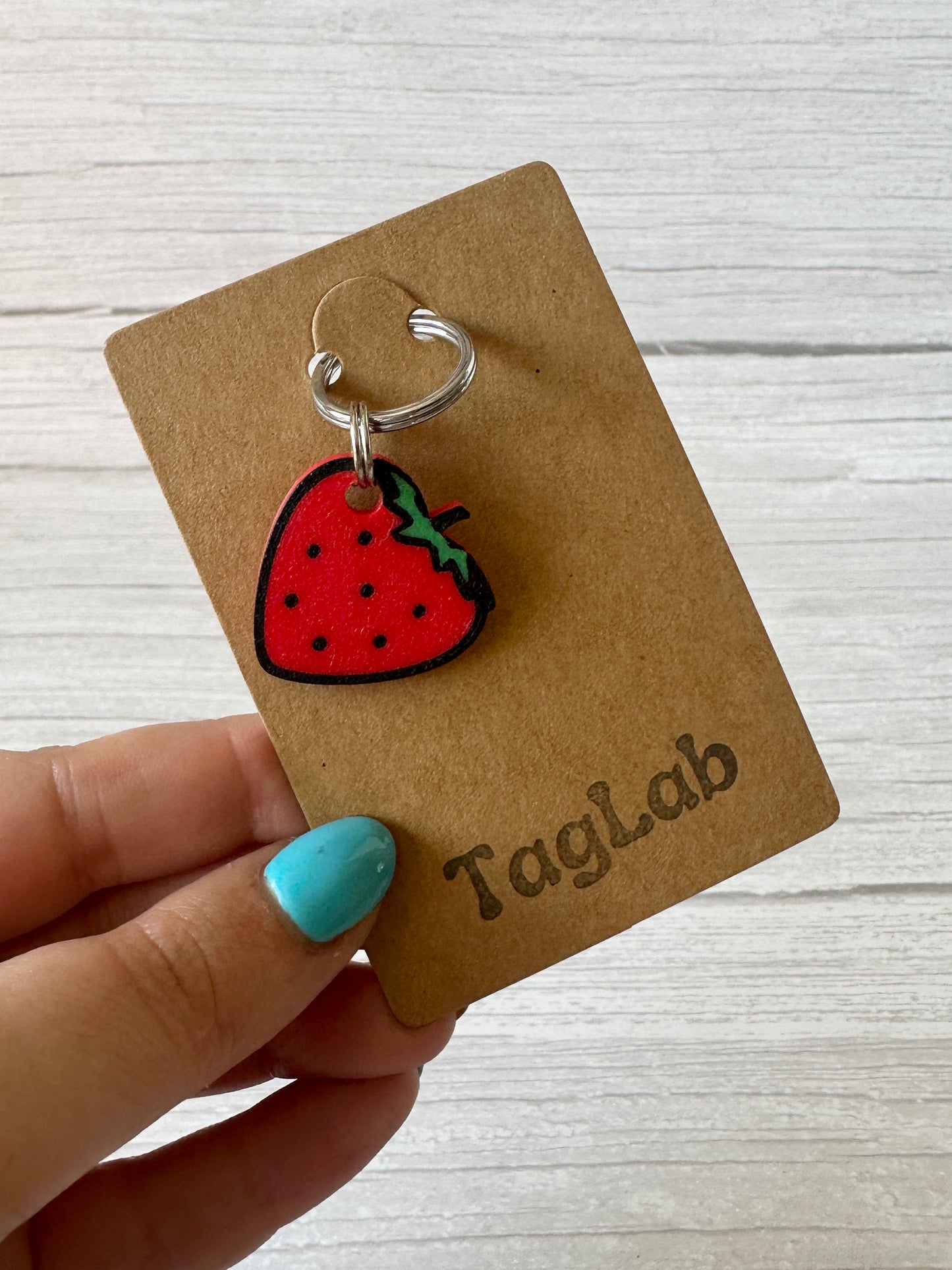 A hand with turquoise nail polish holds a brown "Crafts by Kate" card displaying the Strawberries & Daisies - Arnie & Mac’s Goodies keychain—ideal for brightening your pet bandana. The backdrop is a light wooden surface.