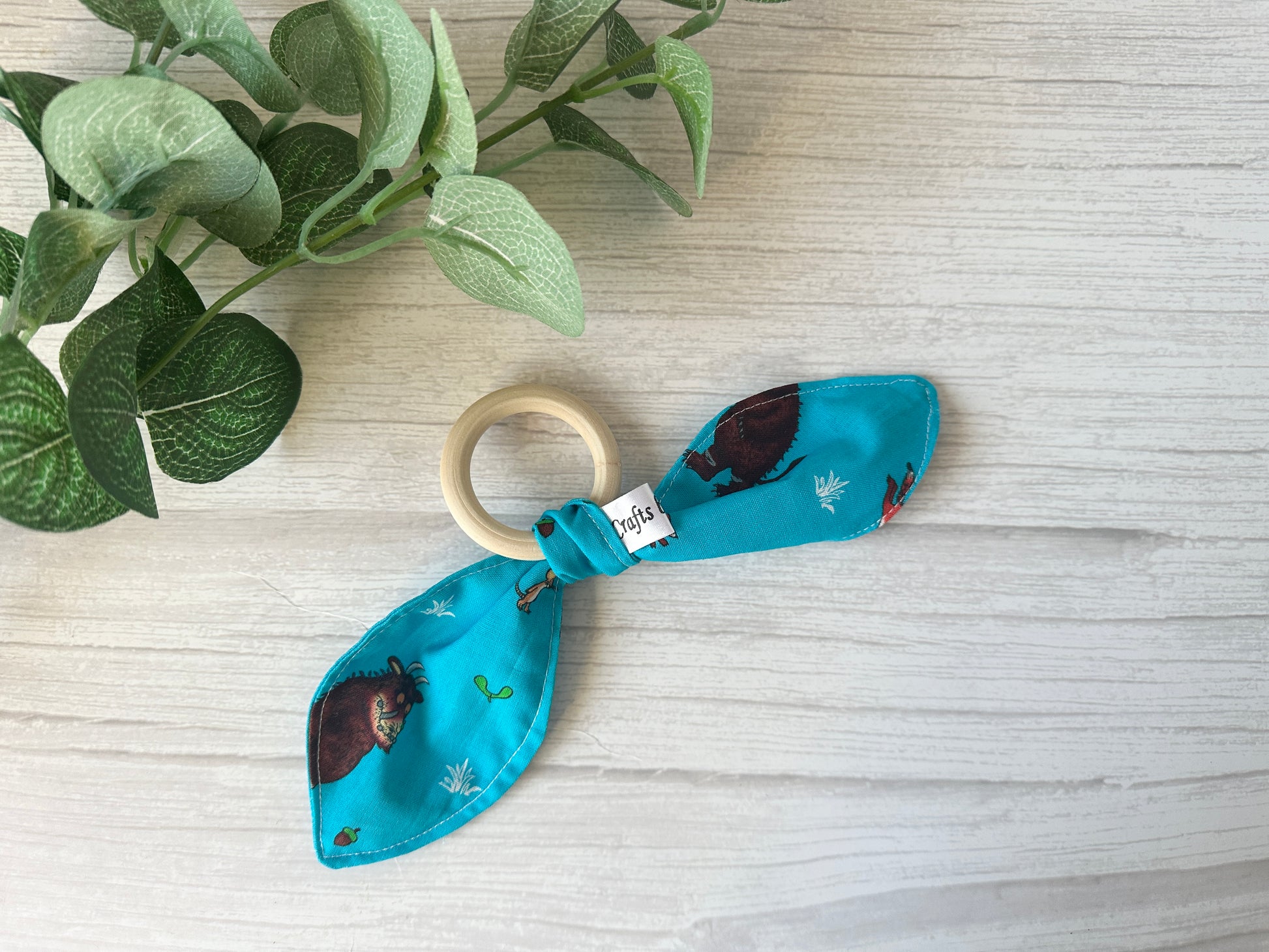 The Cotton & Terry Towelling Baby Gift Set - Gruffalo in blue by Crafts by Kate features a wooden ring with turquoise fabric, playful cartoon animals, and plants. It's perfect for baby gift sets and beautifully presented on a light wooden surface next to a small potted green plant.