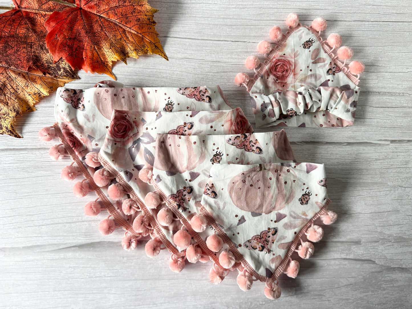 A Cotton Pet Scrunchie Bandana from Crafts by Kate, made using Roses & Pumpkins Fabric and adorned with a floral and pumpkin print, is showcased on a light wooden surface. An autumn leaf in vibrant shades of red and orange is placed beside the bandana, enhancing the fall-themed display.