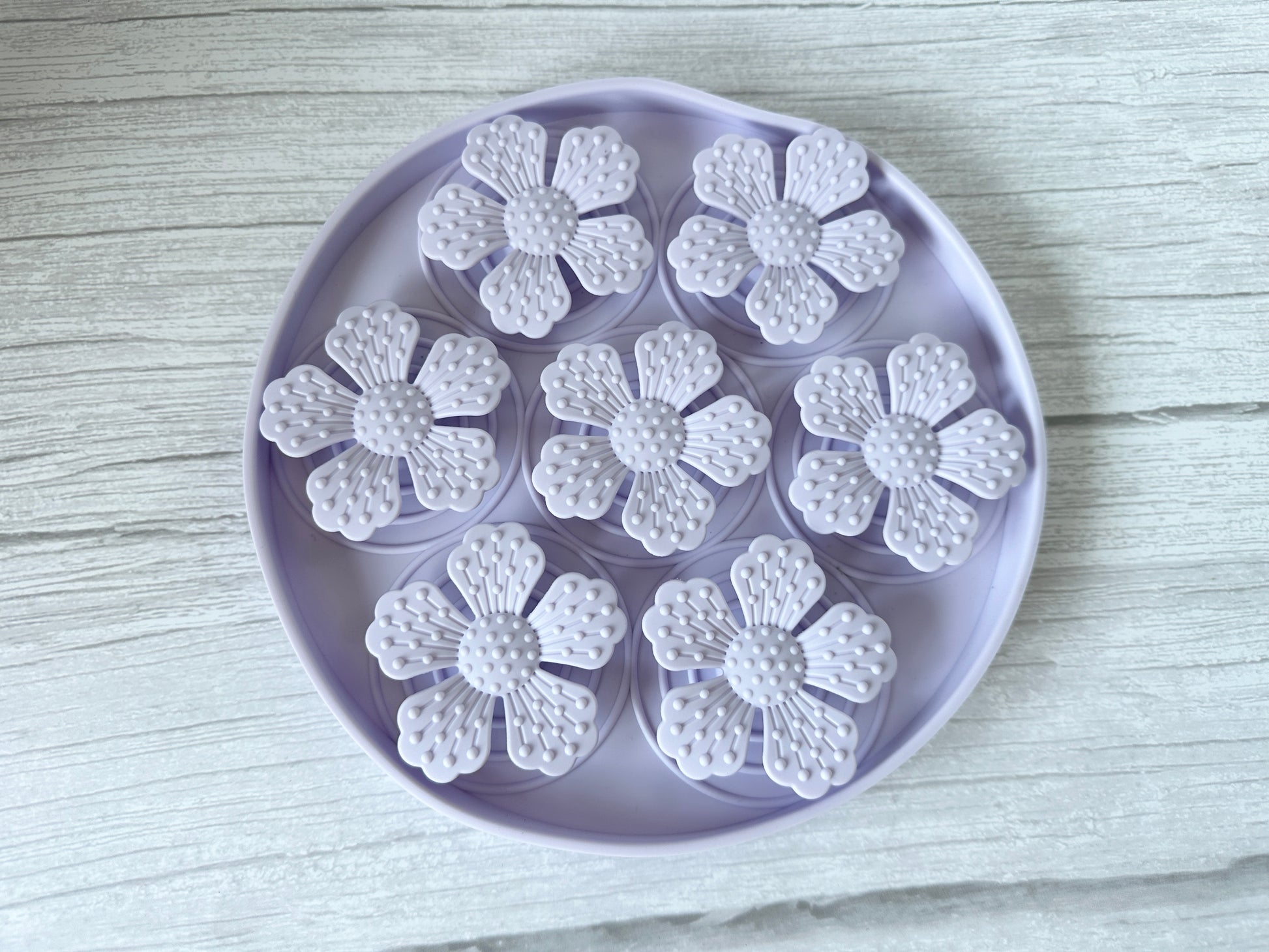 The Crafts by Kate Slow Feeder Pet Mat is a round lavender tray with seven flower-like sections featuring petal designs and suction cups for stability. Placed on a light wooden surface, this washable mat ensures easy cleanup and aids digestion by preventing mealtime messes.