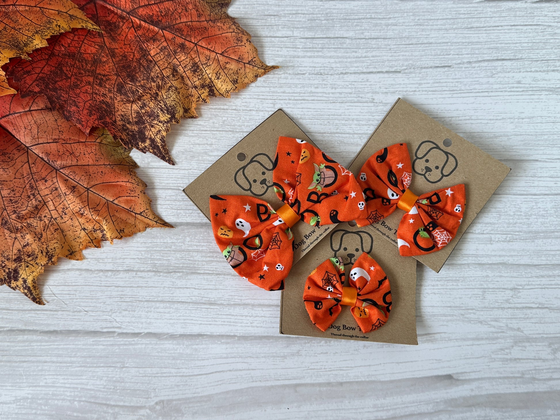 Three Halloween-themed orange bow ties, made by Crafts by Kate, with fun patterns including Baby Yoda fabric, are set on brown cardboard against a light wood surface. Decorated with pumpkins and candy corn, the cotton pet bows have elastic fasteners. Autumn leaves frame the top left corner.