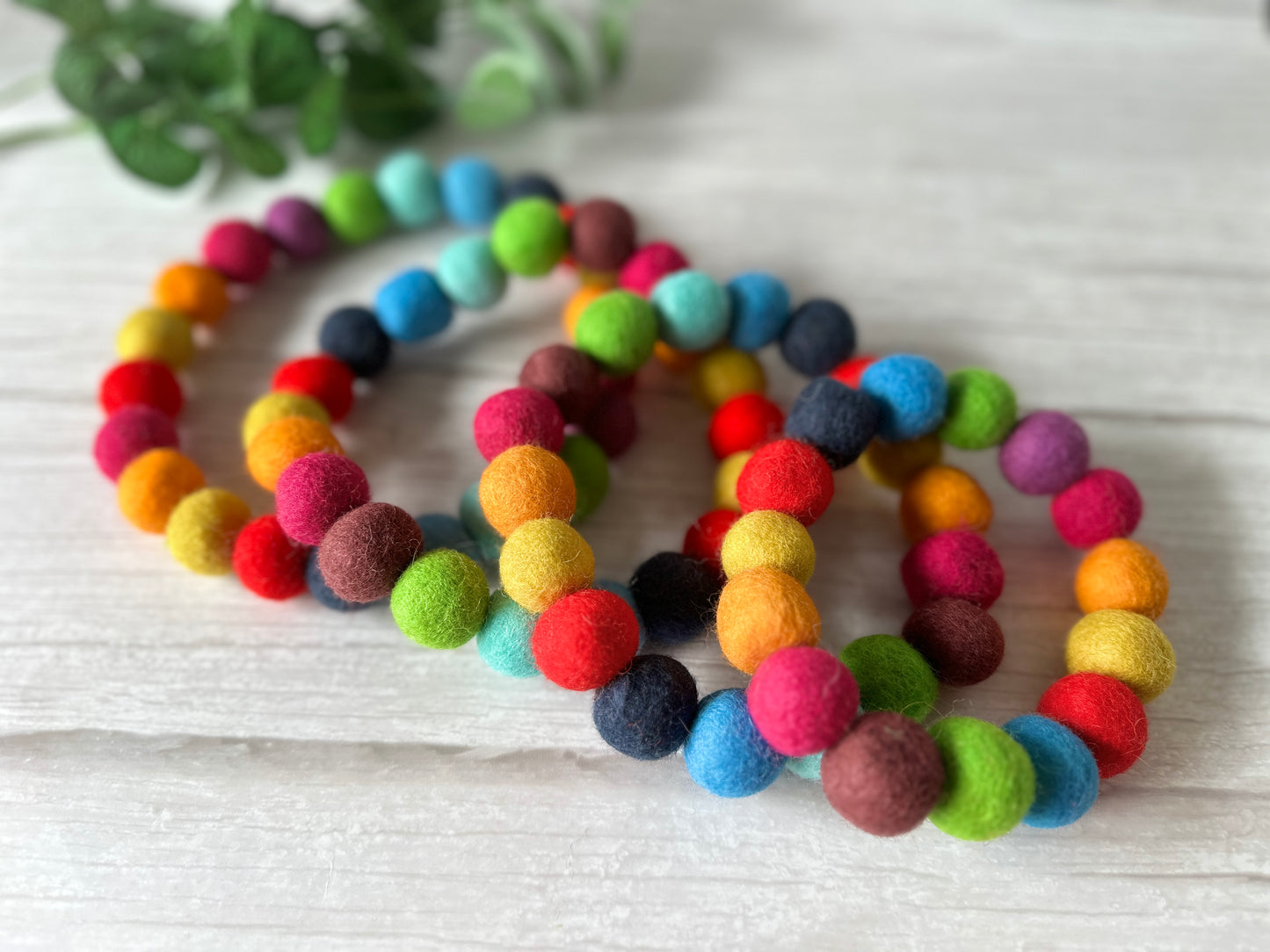 A series of PomPom Pet Collars - Rainbow by Crafts by Kate are laid out on a light wooden surface, each featuring bright and colorful felt pom-poms for a joyful and lively look. In the background, green leaves enhance the natural elegance of this artisanal piece.