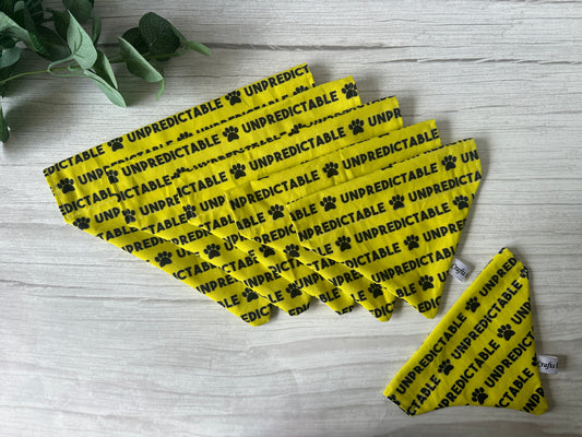 Six yellow “Cotton Pet Bandana with collar attachment - Unpredictable” by Crafts by Kate are fanned on a light wooden surface. Each bandana features black “UNPREDICTABLE” text, paw prints, and a slide-through collar design. A plant sits in the upper left corner.