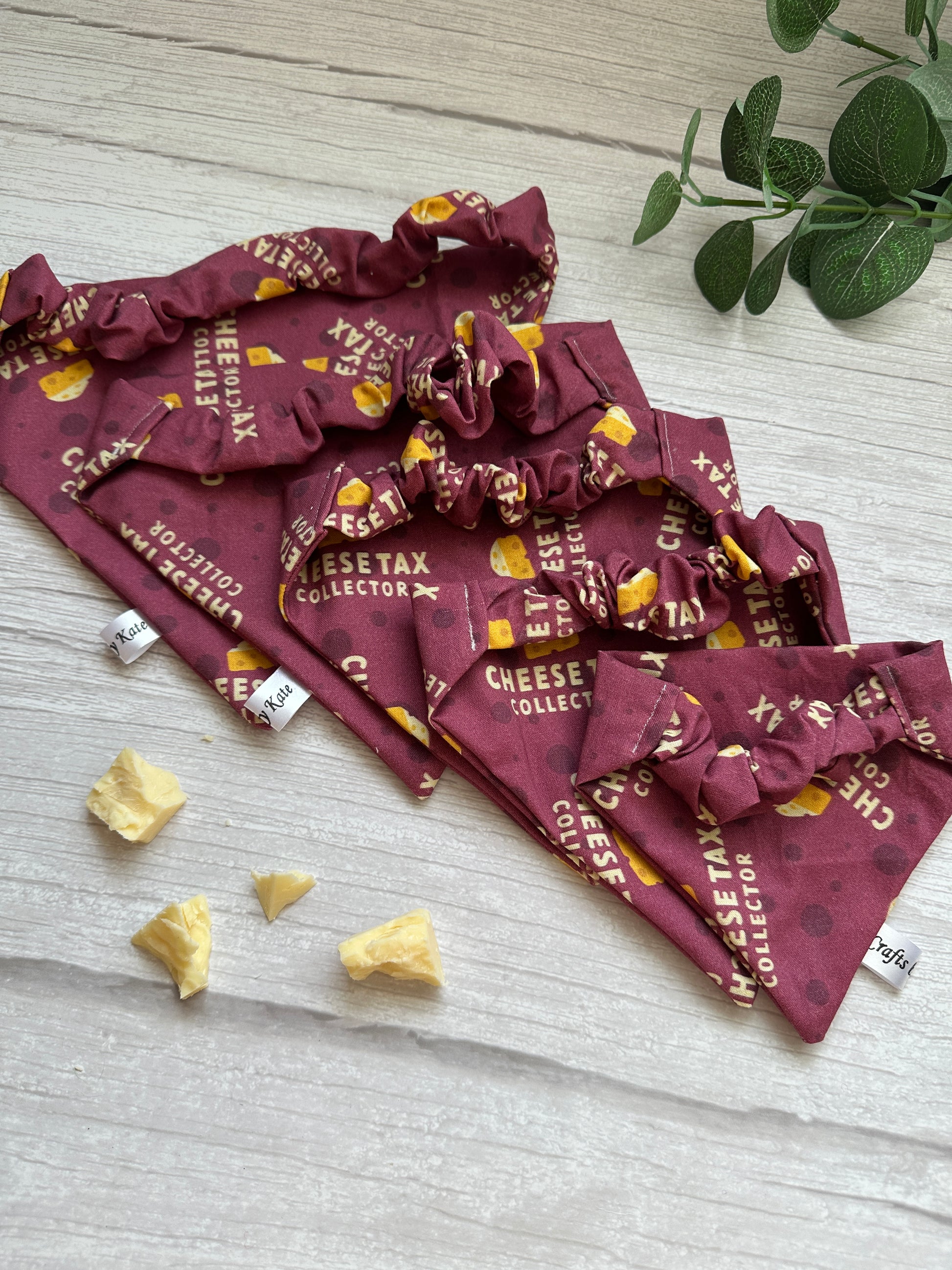 A "Cheesetax Collector" cotton pet scrunchie bandana by Crafts by Kate, featuring cheese illustrations, rests on a wooden surface. Nearby, pieces of cheese and a green plant add to the charming display.