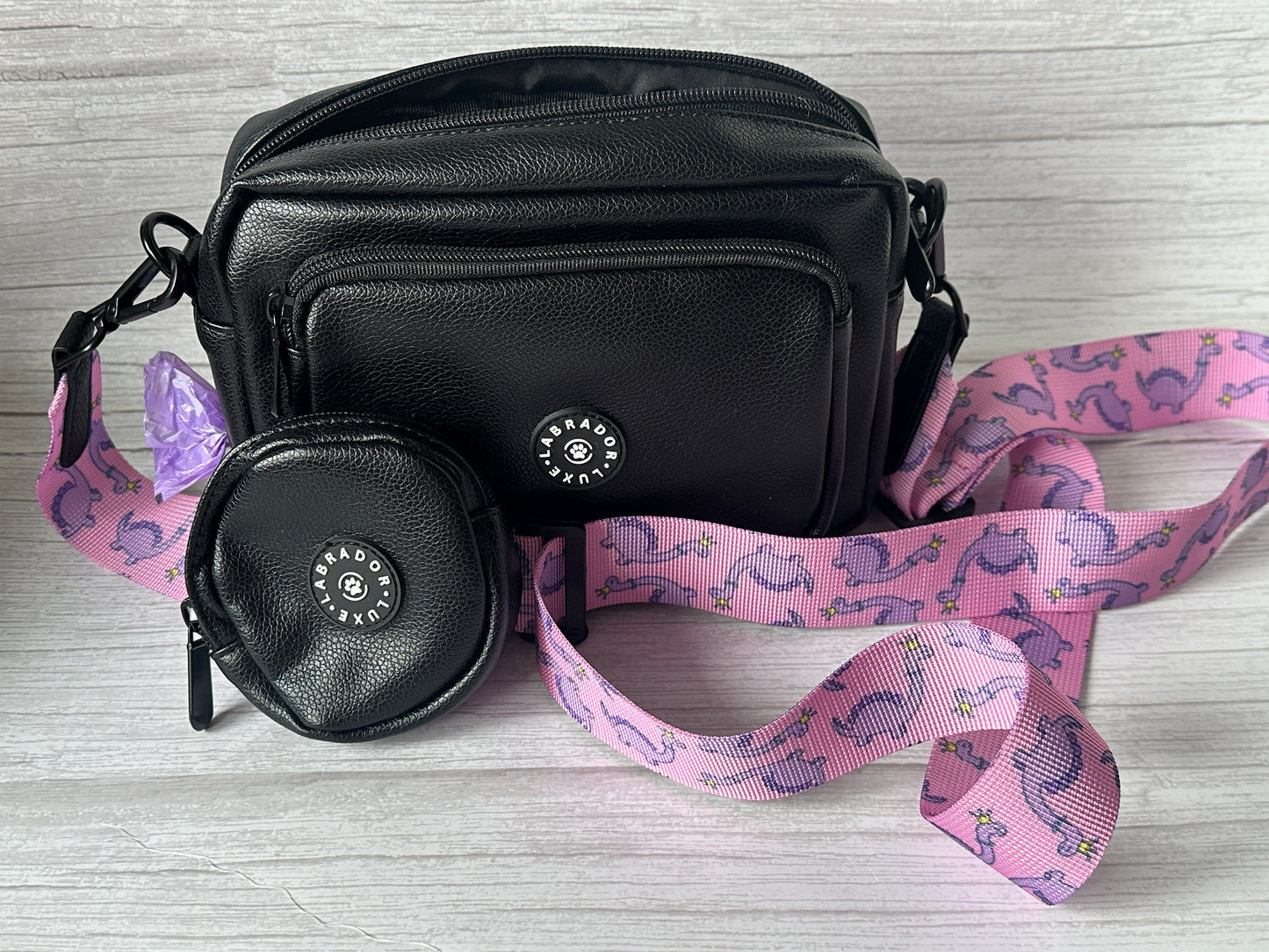 The Crafts by Kate "Millie" Dog Walking Bag, made of small black vegan leather, features a matching round coin pouch and a detachable pink strap with purple dinosaurs and flowers. Both items sit on a light wood surface.