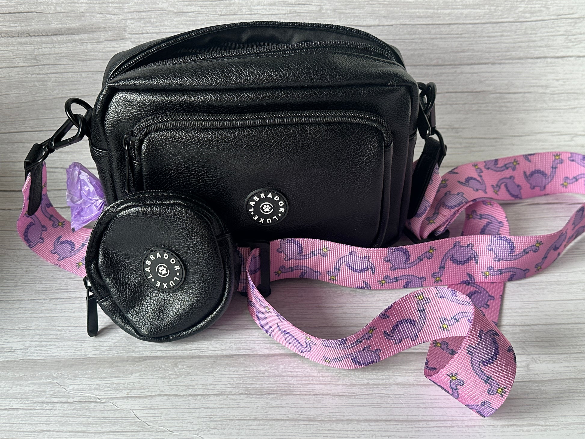 The Crafts by Kate "Millie" Dog Walking Bag, made of small black vegan leather, features a matching round coin pouch and a detachable pink strap with purple dinosaurs and flowers. Both items sit on a light wood surface.