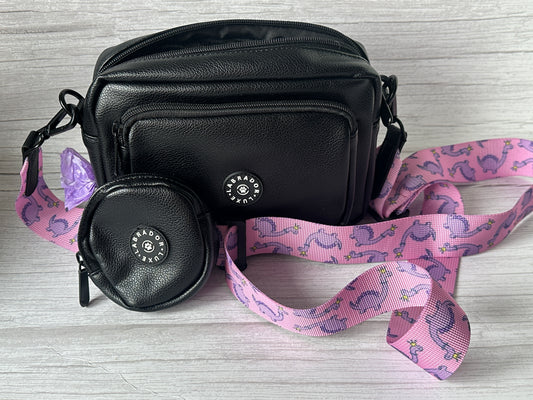 The Crafts by Kate "Millie" Dog Walking Bag, made of small black vegan leather, features a matching round coin pouch and a detachable pink strap with purple dinosaurs and flowers. Both items sit on a light wood surface.