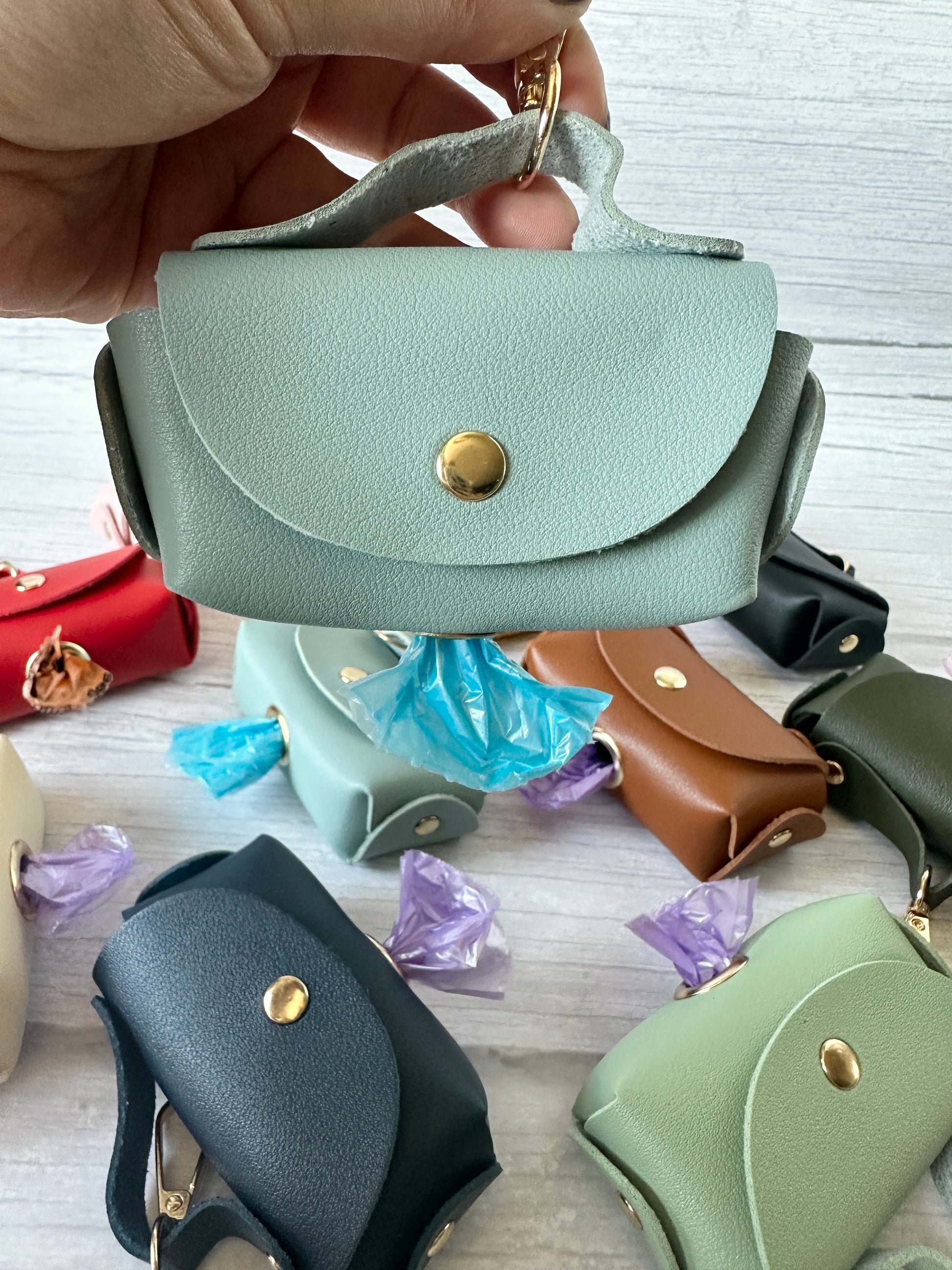 A hand holds a mint green faux leather bag-shaped Pet Poo Bag Holder from Crafts by Kate, featuring a snap button and keyring. Below, other vibrant holders await, showing off brightly colored plastic bags for any adventure.