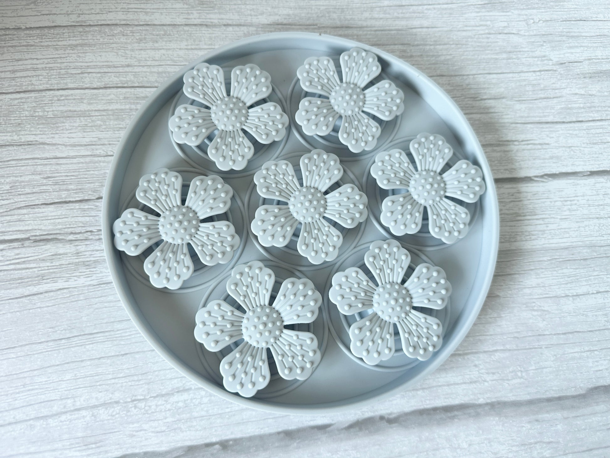 On a light wooden surface, the Crafts by Kate Slow Feeder Pet Mat showcases seven white flower-shaped objects, each with five petals and textured centers that resemble tiny suction cups.