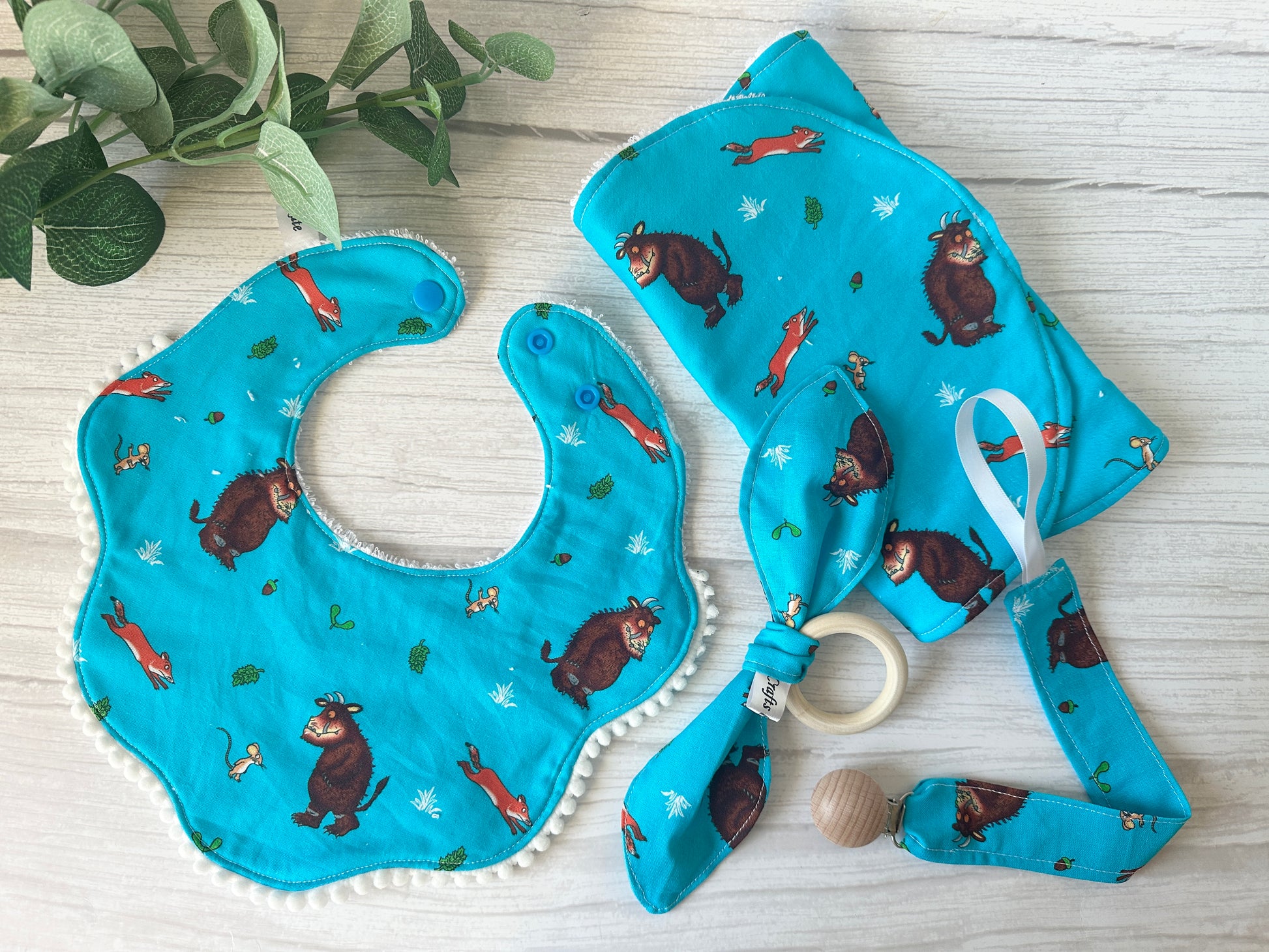 The "Cotton & Terry Towelling Baby Gift Set - Gruffalo in blue" by Crafts by Kate, featuring a bib, teething ring, and pacifier clip with cartoon fox and bear patterns on ultra-soft cotton, sits charmingly on wood beside a small plant.